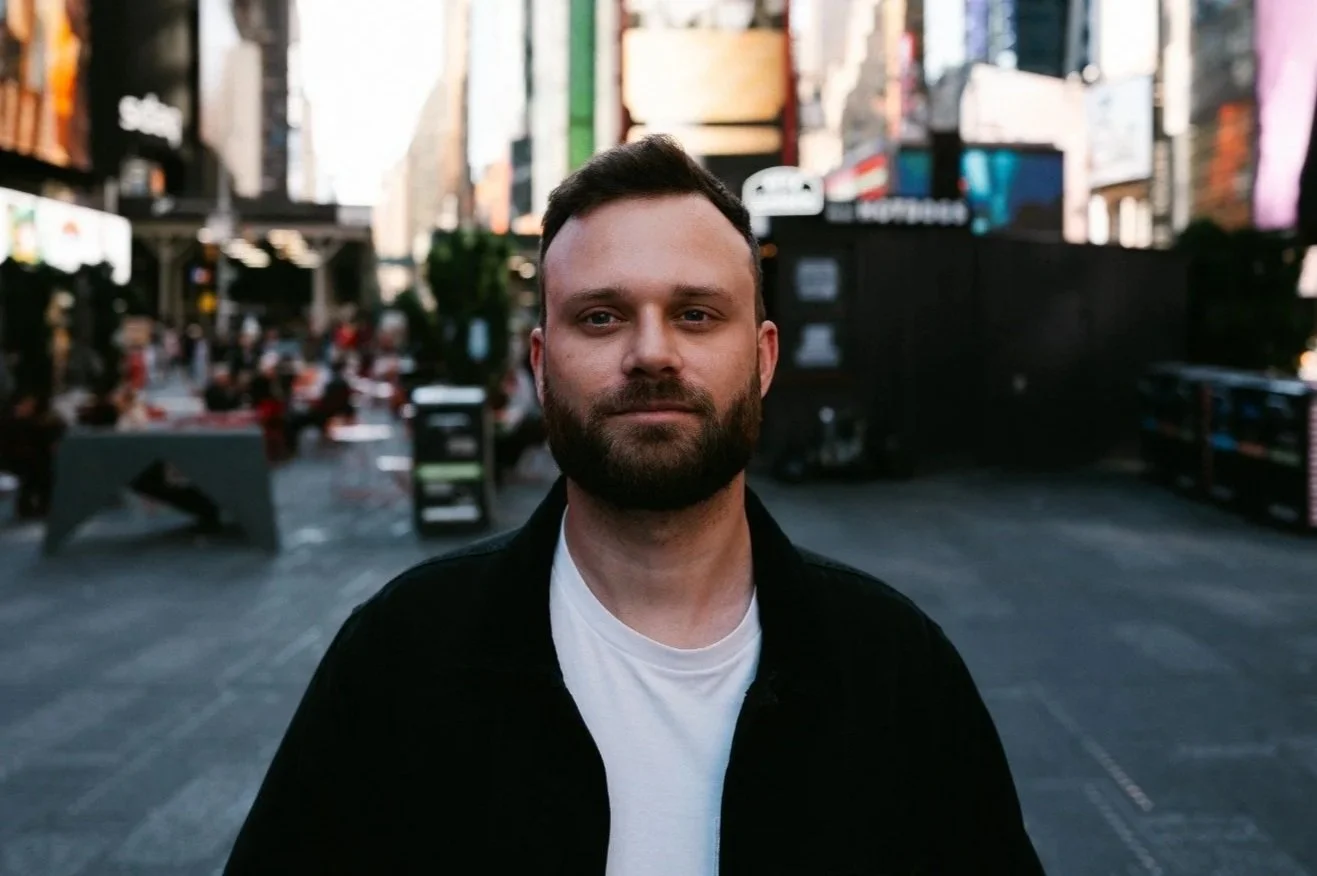 A man with a beard and short hair standing in a city square with buildings and digital billboards in the background.