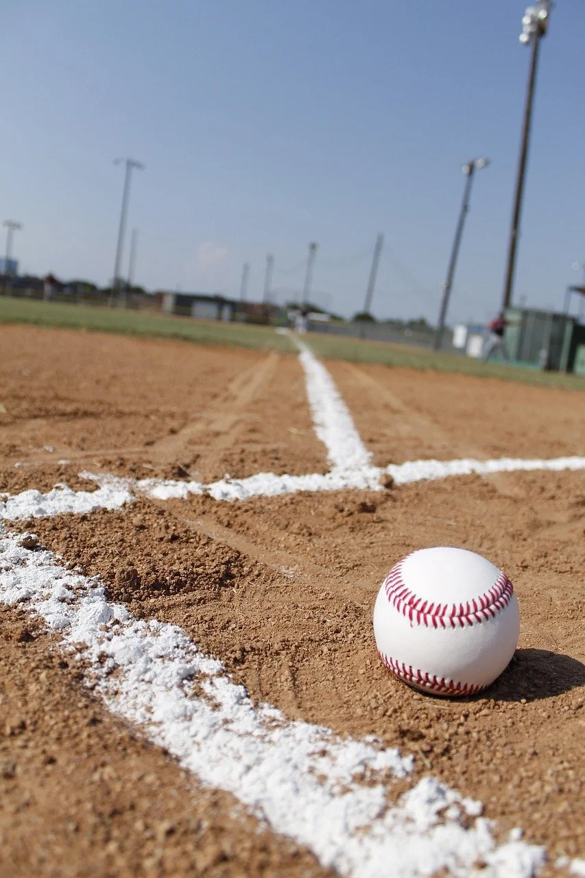 Close-up of a baseball resting on the dirt near the white chalk line on a baseball field, with the outfield, field lights, and blue sky in the background.