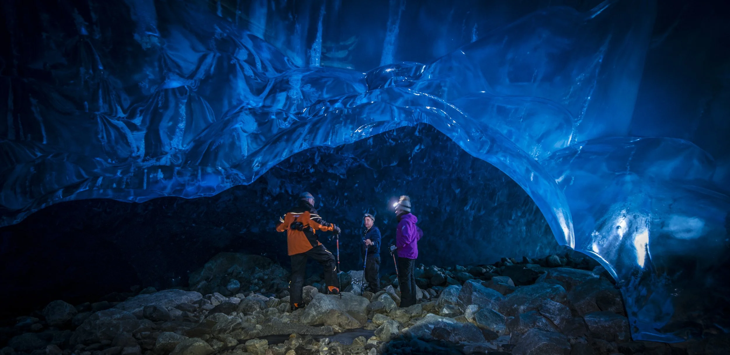 Three adventurers in winter gear exploring an ice cave illuminated with blue light, standing on rocky terrain.