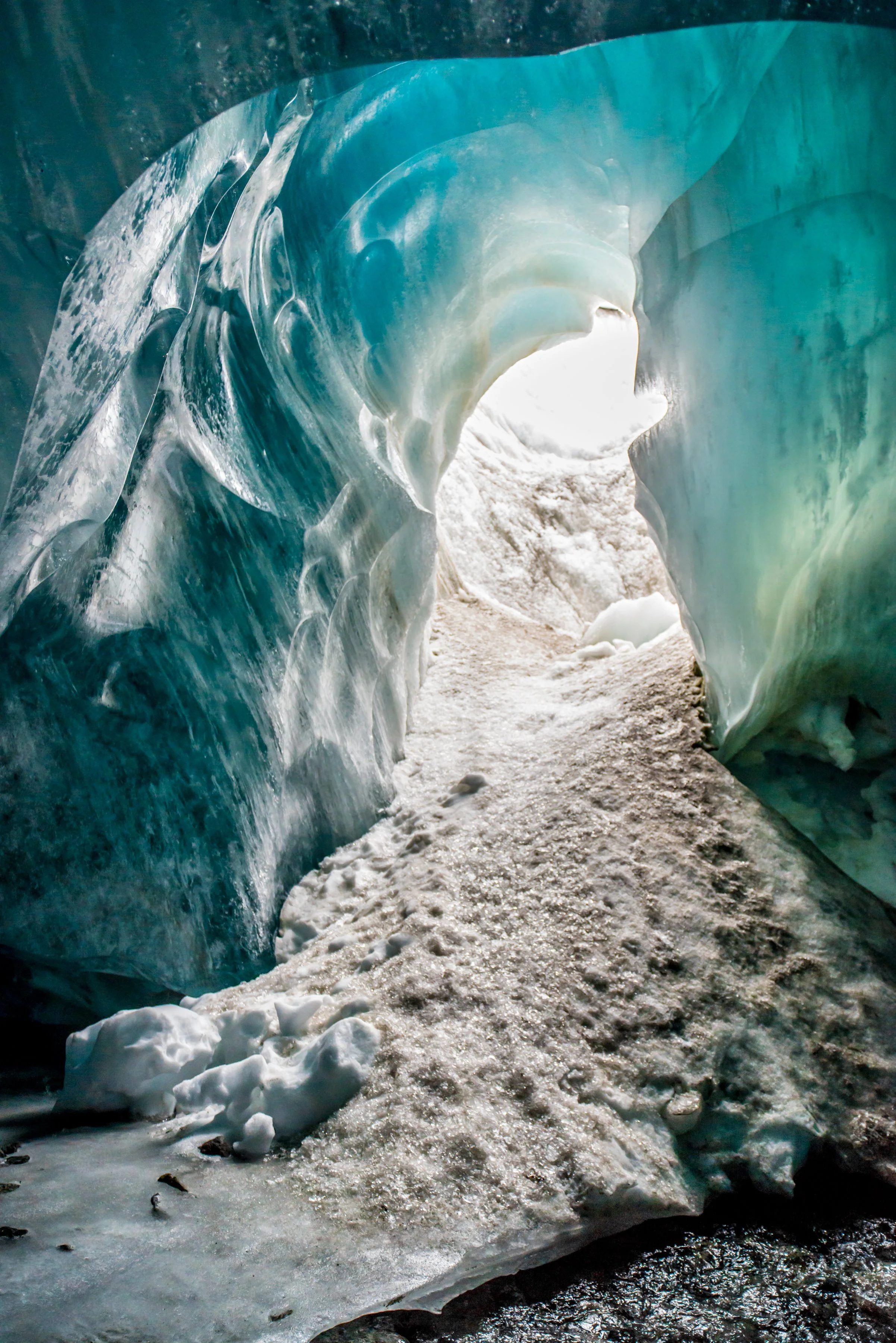 Inside a glacier ice cave with blue translucent ice walls and a snow-covered floor, with light coming from the opening ahead.