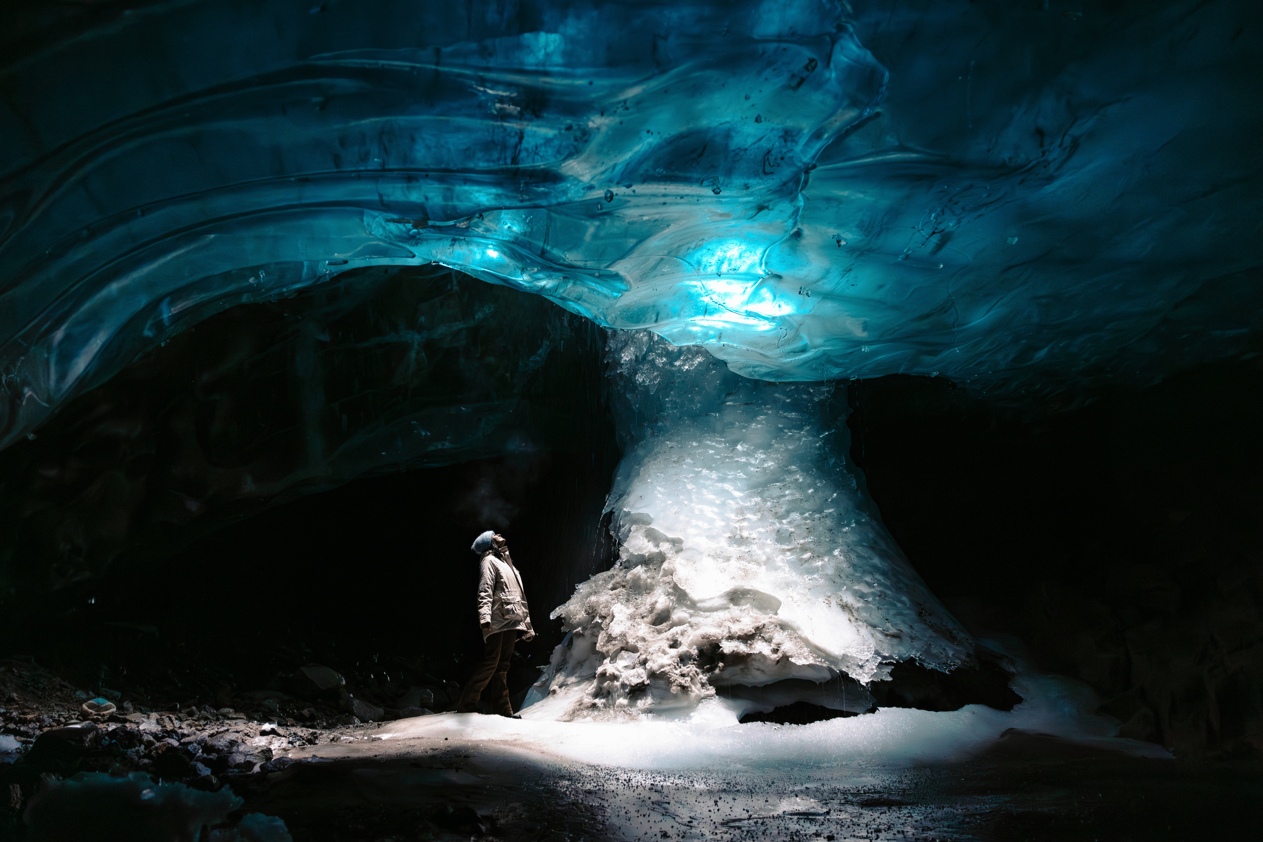 A person standing inside an ice cave with ice formations on the ceiling and walls, and light glowing from the opening.