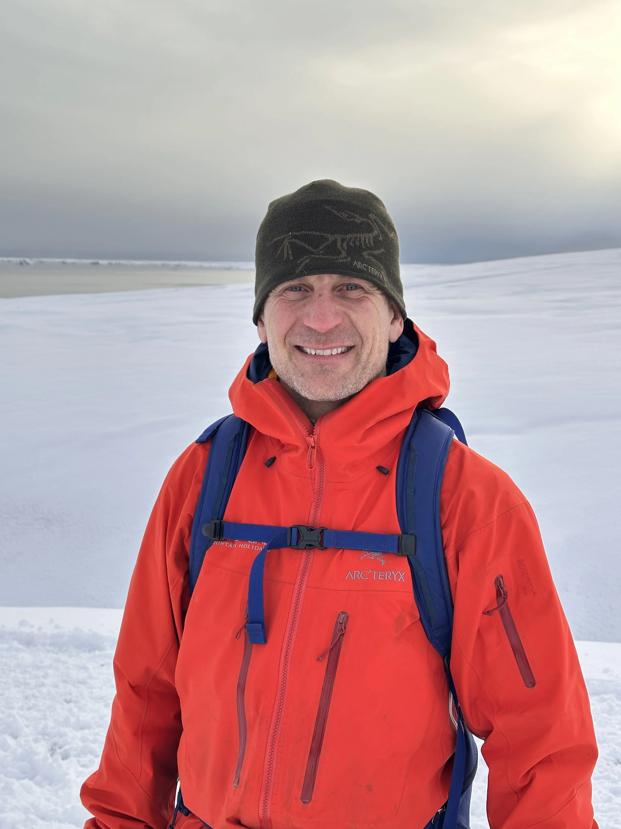 A smiling man in a red winter jacket and a black beanie standing in a snowy landscape with a cloudy sky in the background.