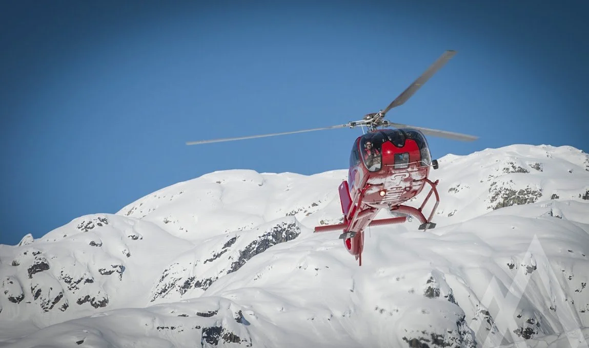A red helicopter flying over snowy mountain peaks during the day.