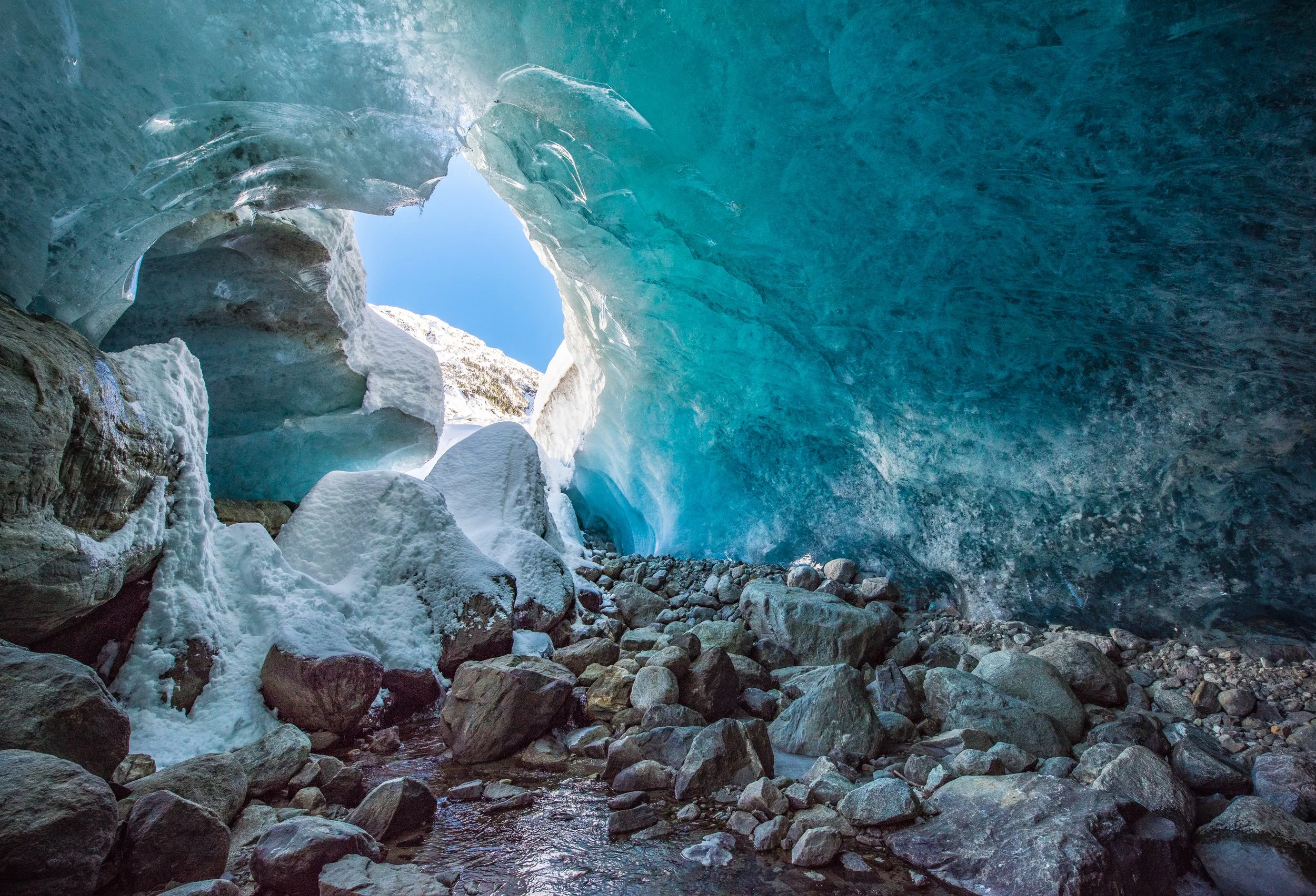 An ice cave with a view of blue ice arch and snow-covered rocks inside, with a small stream flowing through the rocks.