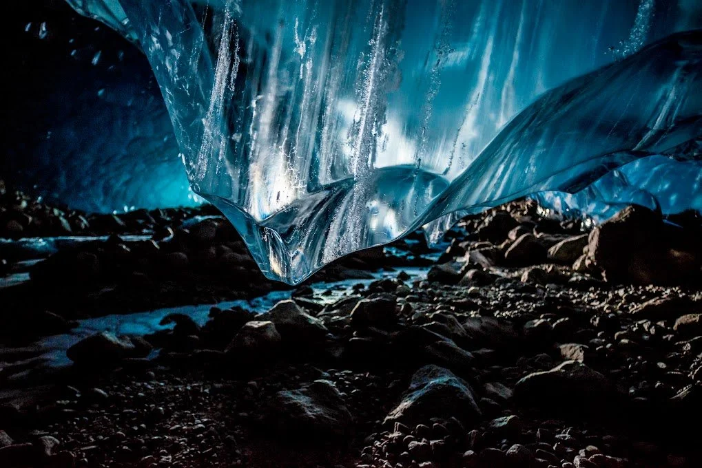 Close-up of a large ice formation or ice cave with sunlight reflecting off the smooth ice surface, with rocks and small stones on the ground.