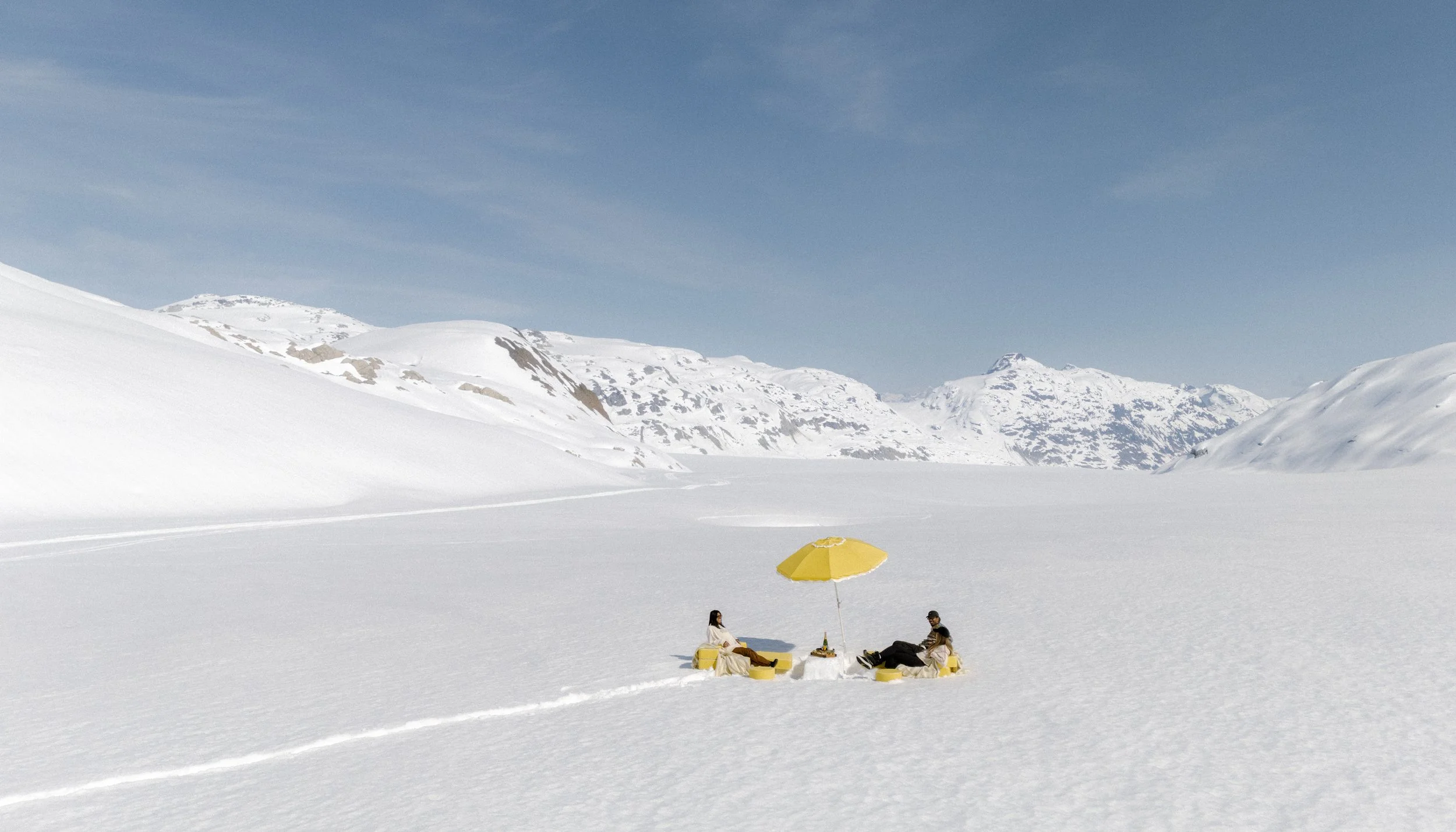 Two people sitting on yellow lounge chairs under a yellow umbrella on snow-covered landscape with mountains in the background.