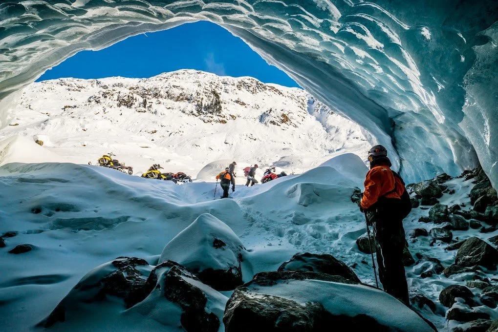 People in winter gear exploring an icy cave with snow-covered mountains outside.
