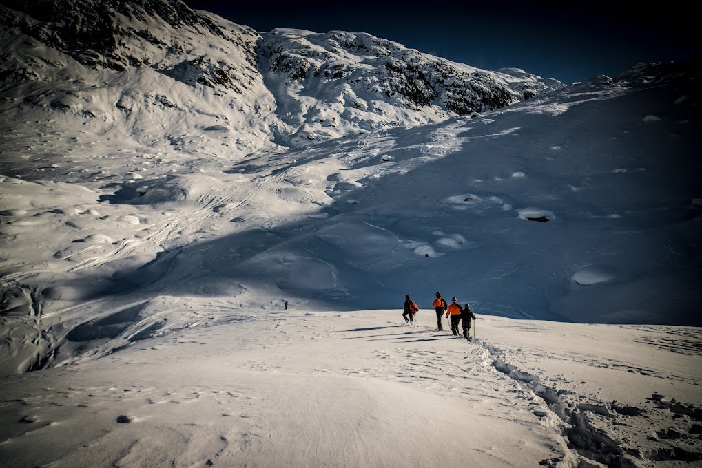 Four people hiking in a snow-covered mountain landscape, with rugged terrain and snowdrifts.