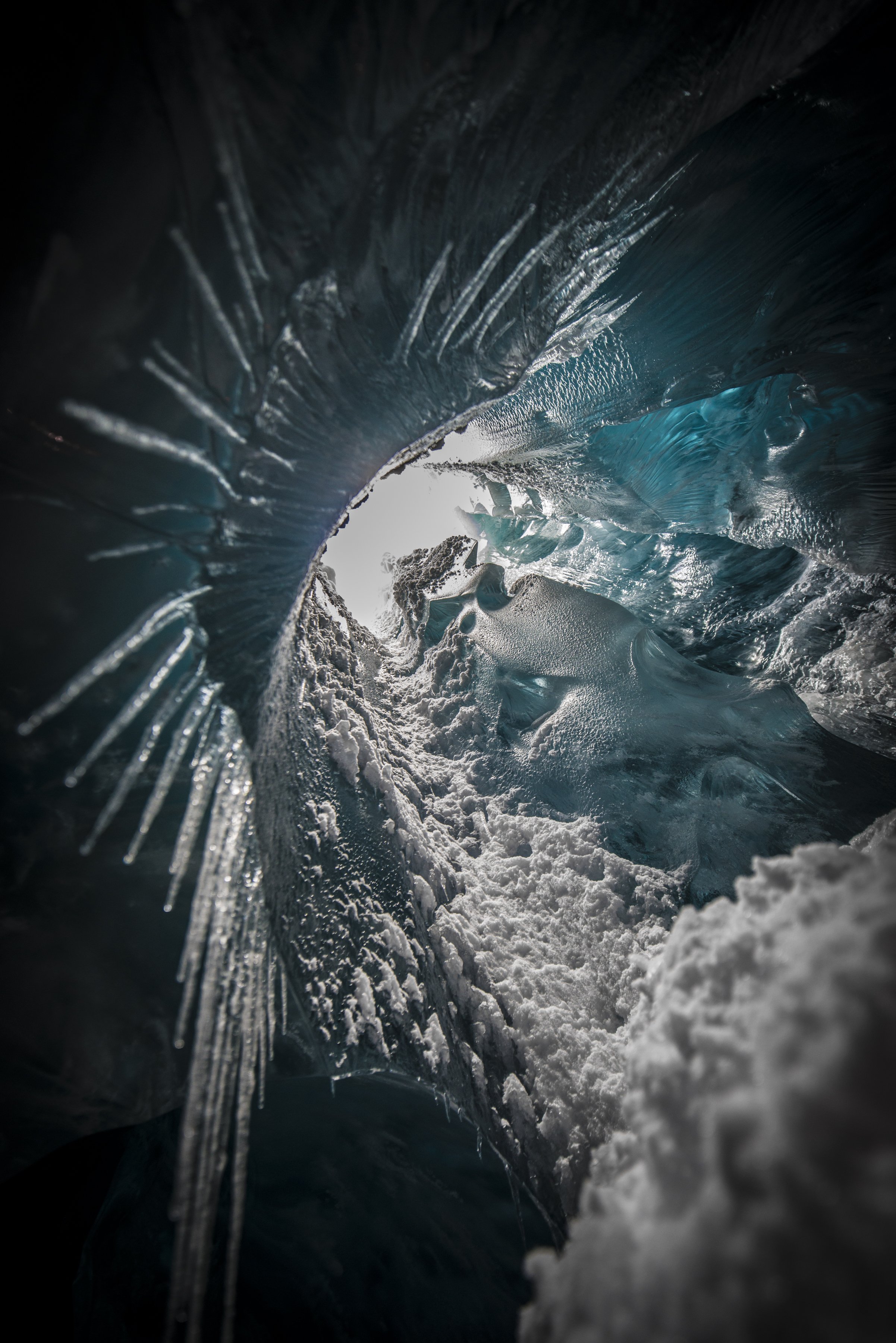 Inside a glacier's icy tunnel showing blue ice and snow with light coming from the opening.