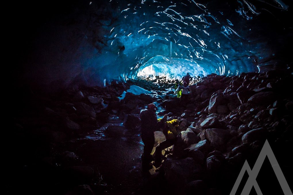 People exploring inside a glowing ice cave with blue and white light, surrounded by rocks.