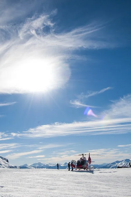 Helicopter on snow-covered landscape with mountains in the background and a bright sun in the sky.
