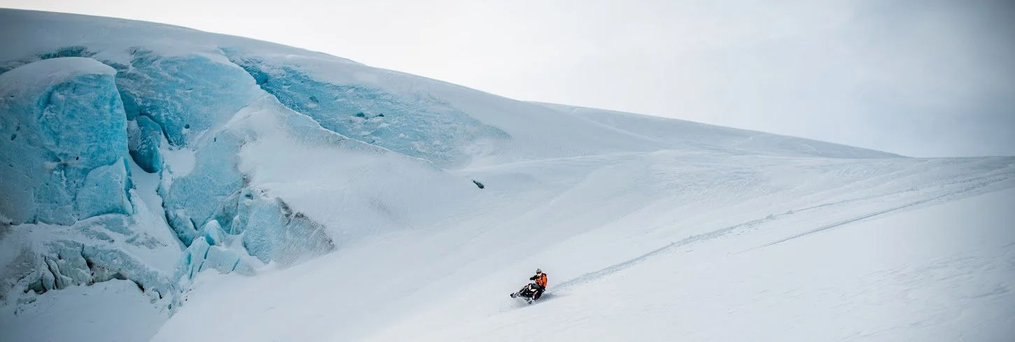 A person riding a snowmobile on a snow-covered slope near a large glacier.