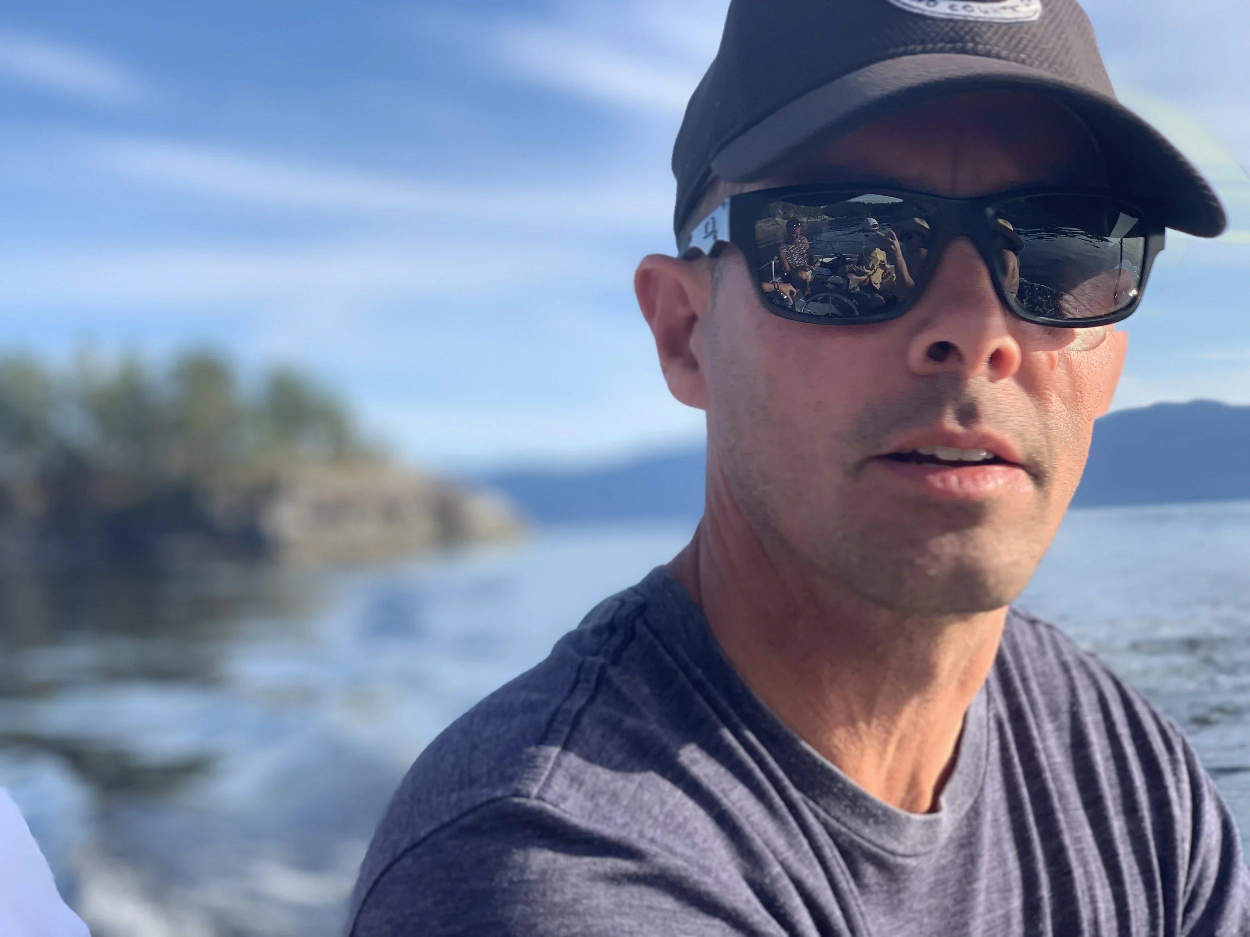 A man wearing sunglasses and a baseball cap, on a boat, with water and a small island with trees in the background, during daytime.