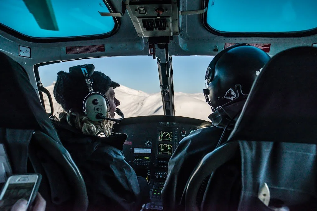 View of the cockpit of a helicopter with two pilots wearing helmets, flying over a snow-covered mountainous landscape.