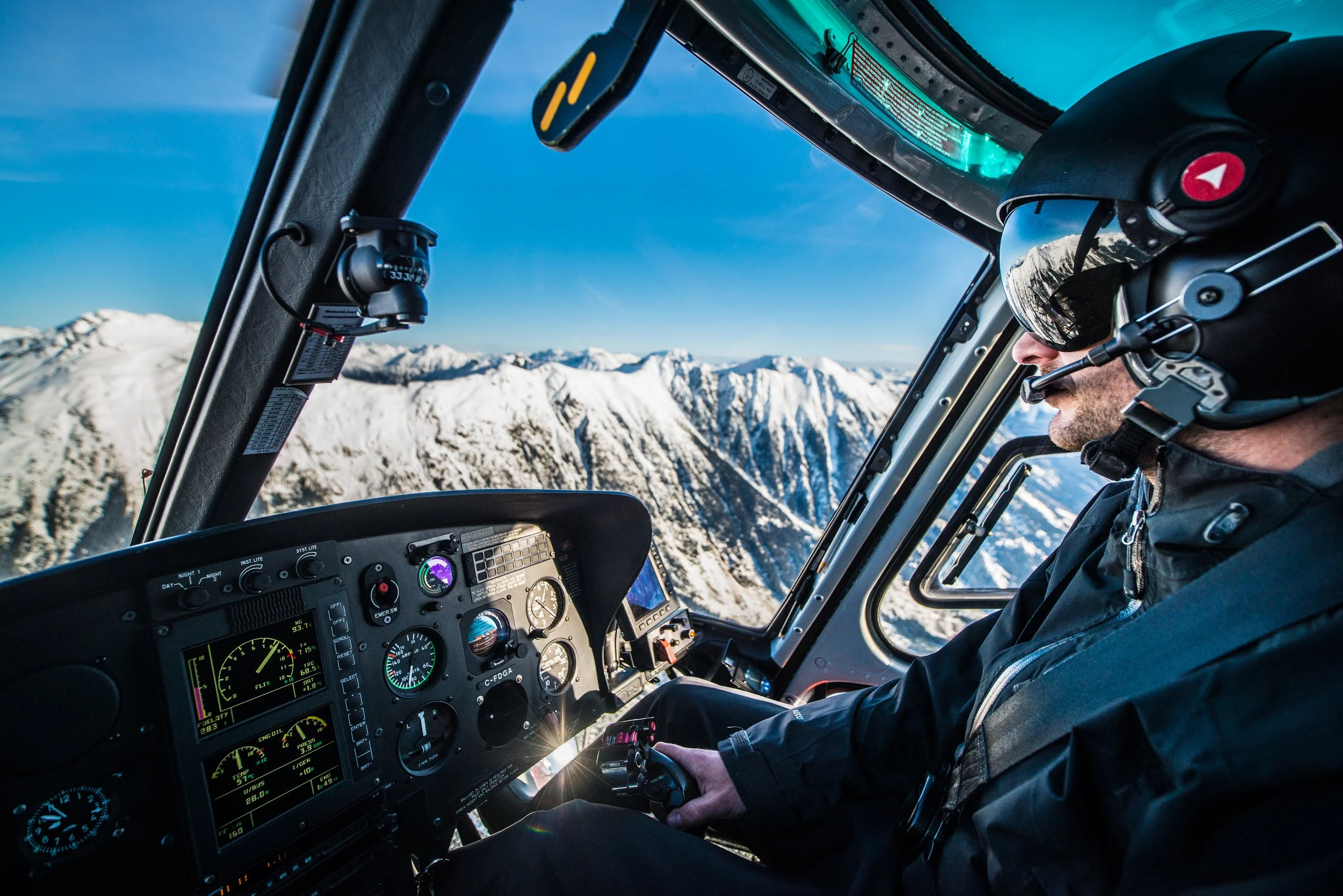 A helicopter pilot operating the controls inside a helicopter cockpit with a view of snow-covered mountains outside.