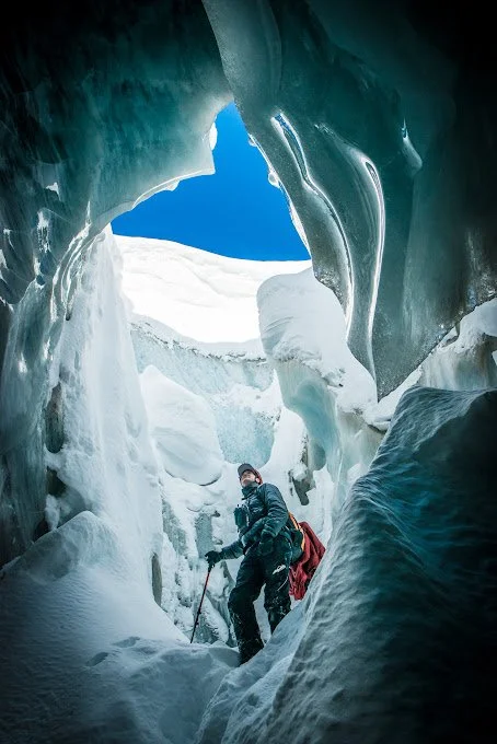 Person exploring an icy glacier cave with snow and ice formations and a blue sky visible overhead.