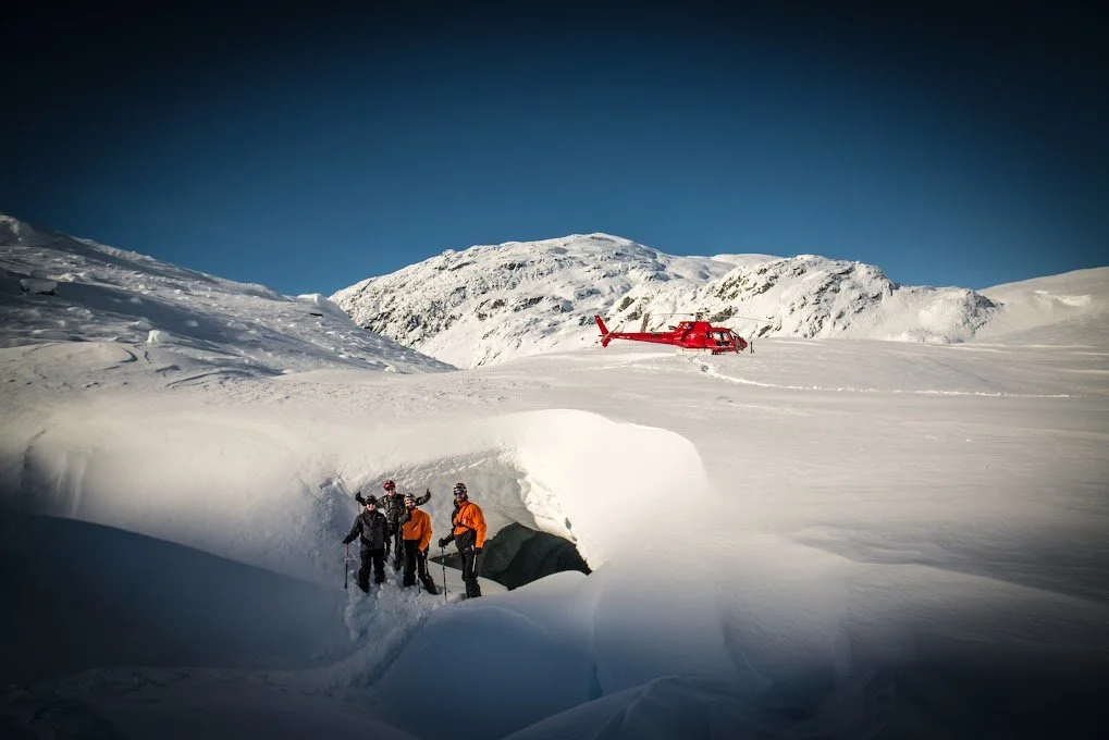 A team of four outdoor explorers in winter gear near an ice cave entrance in a snowy landscape with a red helicopter on a hill in the background.