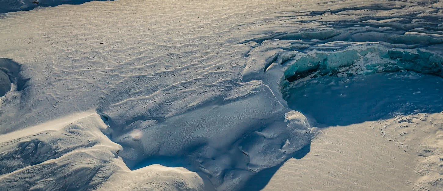 Close-up of a snow and ice-covered landscape with a crevasse or ice fissure in the snow showing blue ice underneath.