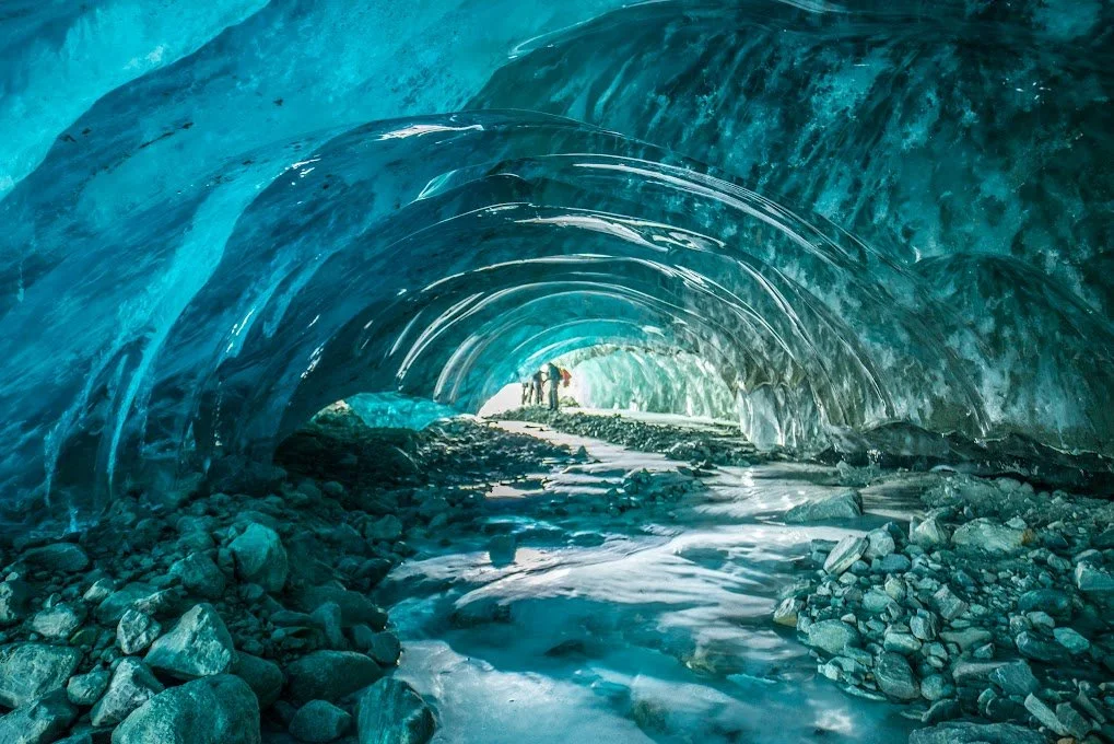 People walking inside an ice cave with blue ice walls and rocky ground.