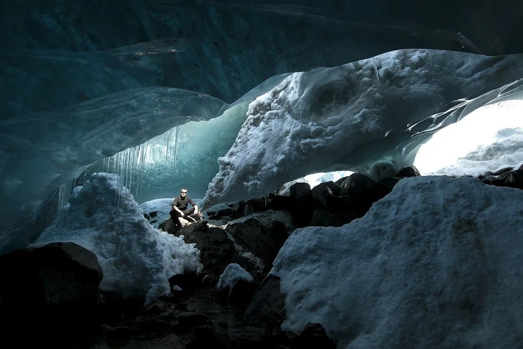 A person sitting among ice and snow inside a glacier with blue ice formations and icicles hanging from the ceiling.