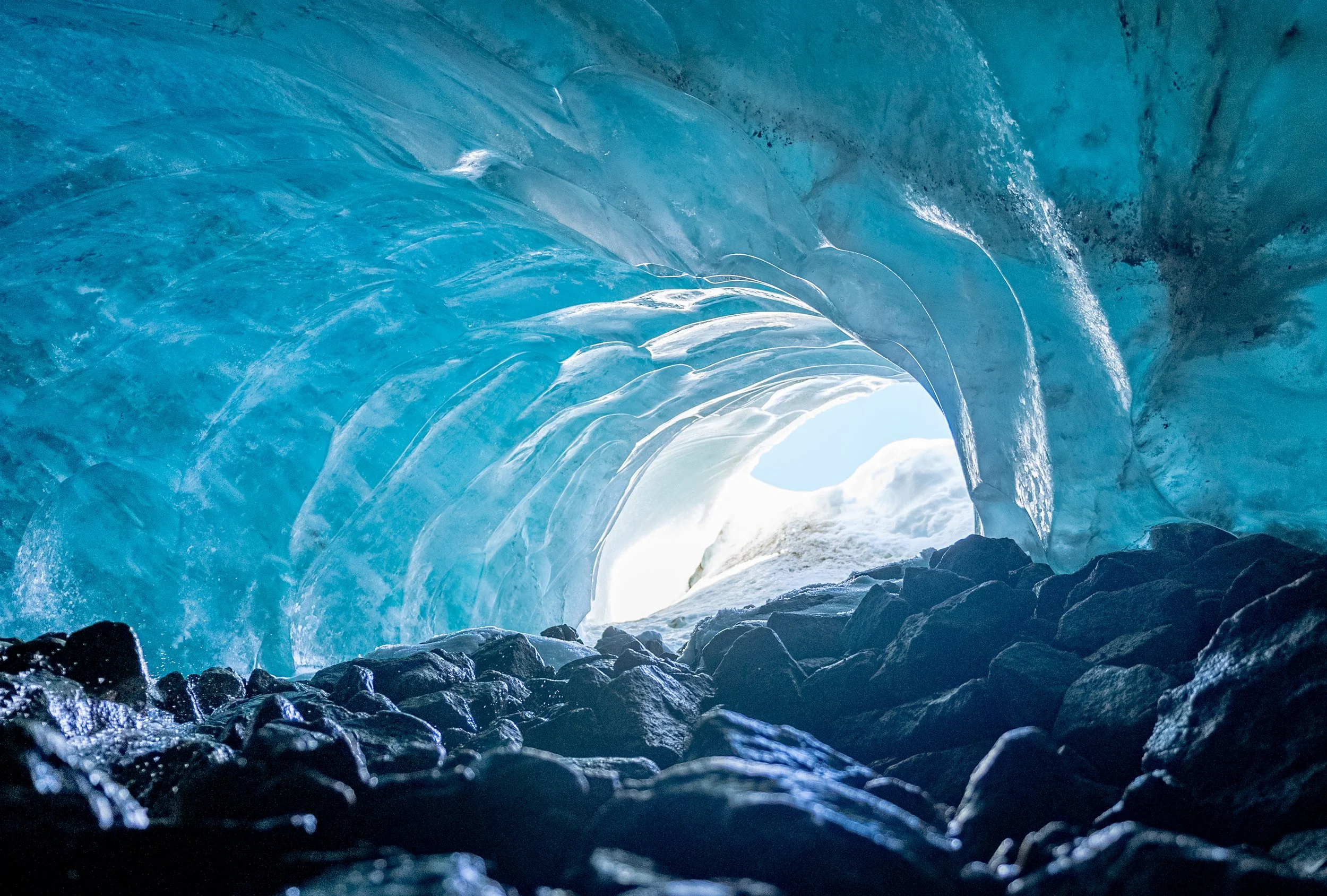 Inside an icy glacier cave with blue ice formations and a view of the bright outside through the cave opening, with dark rocks at the bottom.