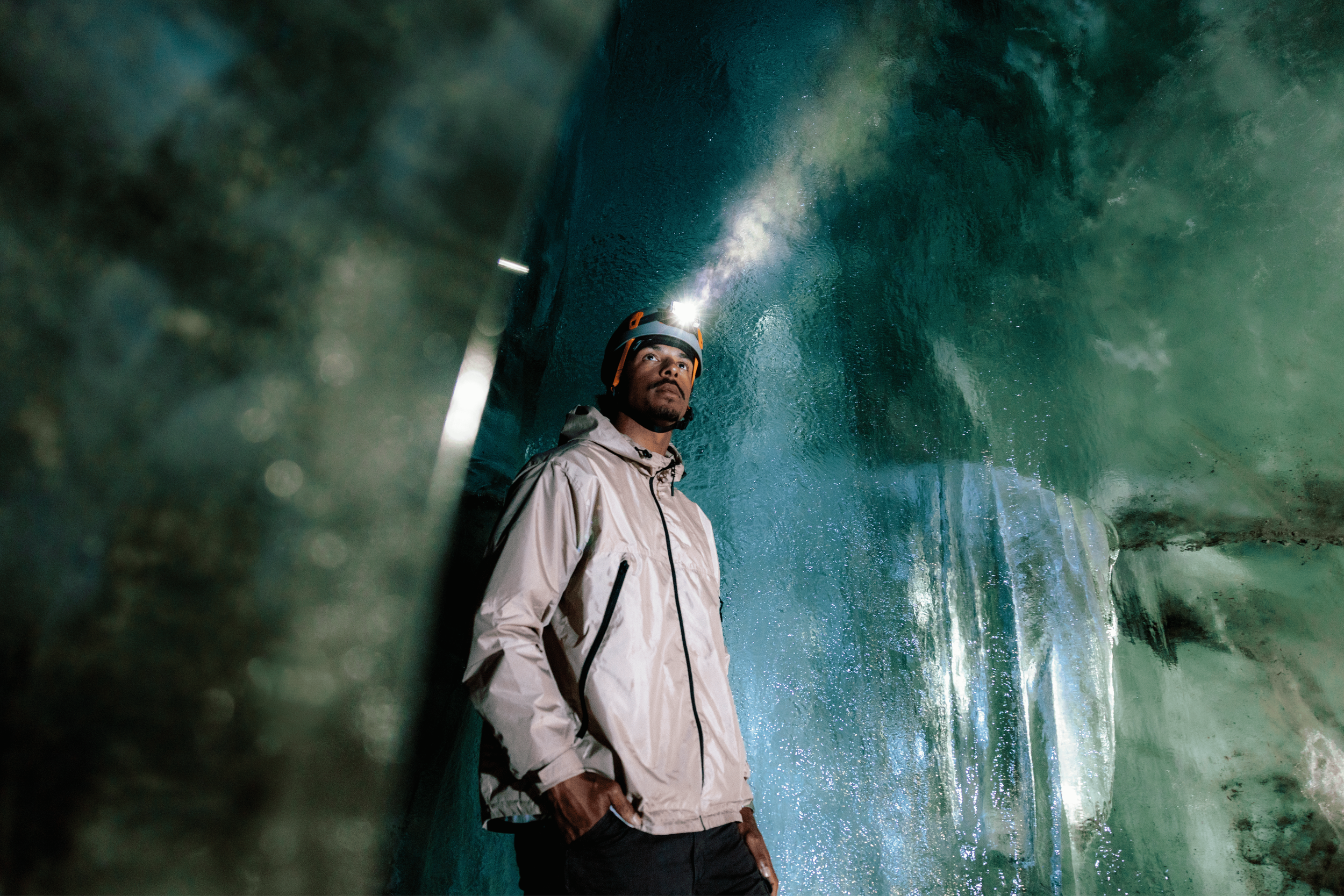 Person wearing an orange helmet and a beige jacket standing inside a large cave with water.