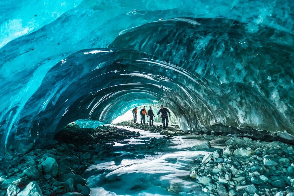Group of people walking inside a large ice cave with blue ice formations and rocky floor.