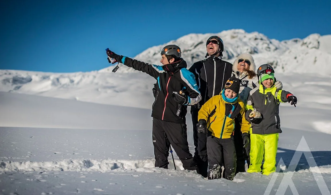 Group of five people in winter clothing and helmets smiling and standing in the snow in front of snow-covered mountains.