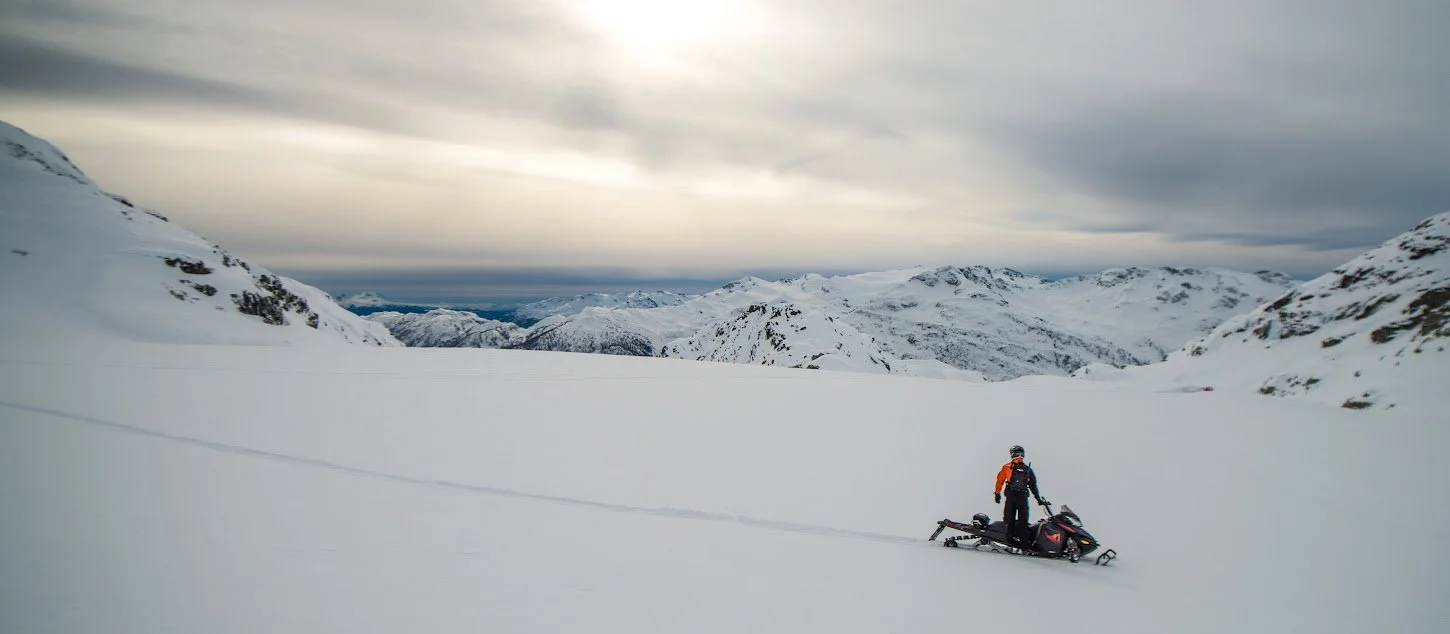 A snowmobile rider in winter gear standing beside a snowmobile on a snowy landscape with snow-covered mountains and cloudy sky in the background.