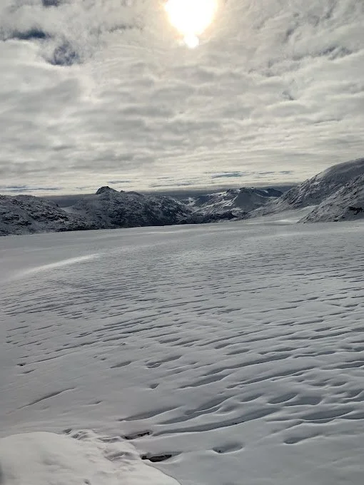 Snow-covered landscape with mountains in the distance under cloudy sky, and the sun partially obscured by clouds.