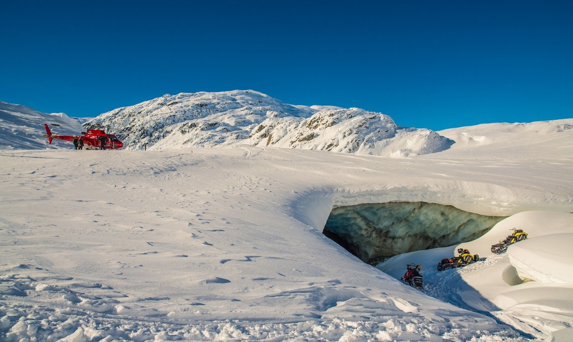 A red helicopter parked on snow-covered terrain in a cold, icy landscape with snow-covered mountains in the background. Several snowmobiles are visible near a large ice crevasse.