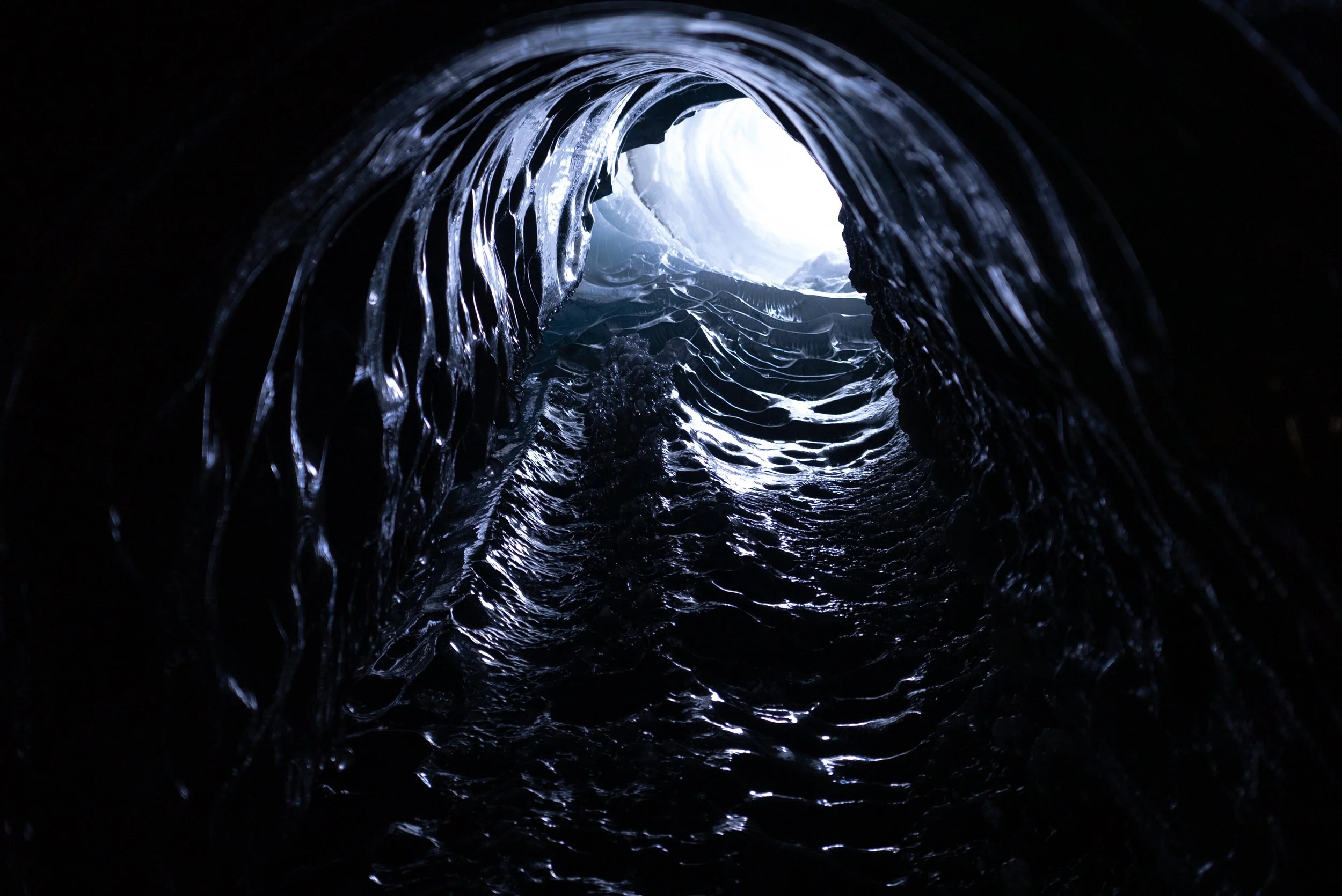 Looking up through a dark, icy cave towards the opening with bright light illuminating the ice formations and water inside.