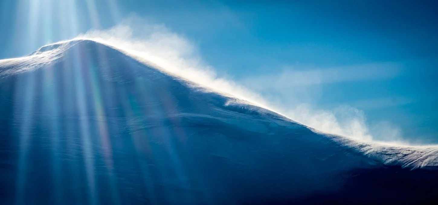 Close-up of a snow-covered mountain peak with sunlight shining on it and a clear blue sky in the background.