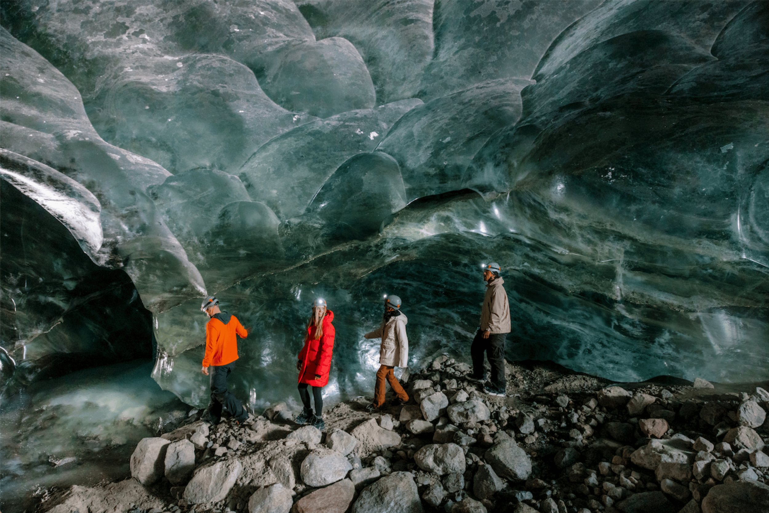 Group of four people in winter clothing and helmets exploring inside a glacier with icy, translucent blue walls and rocky floor.