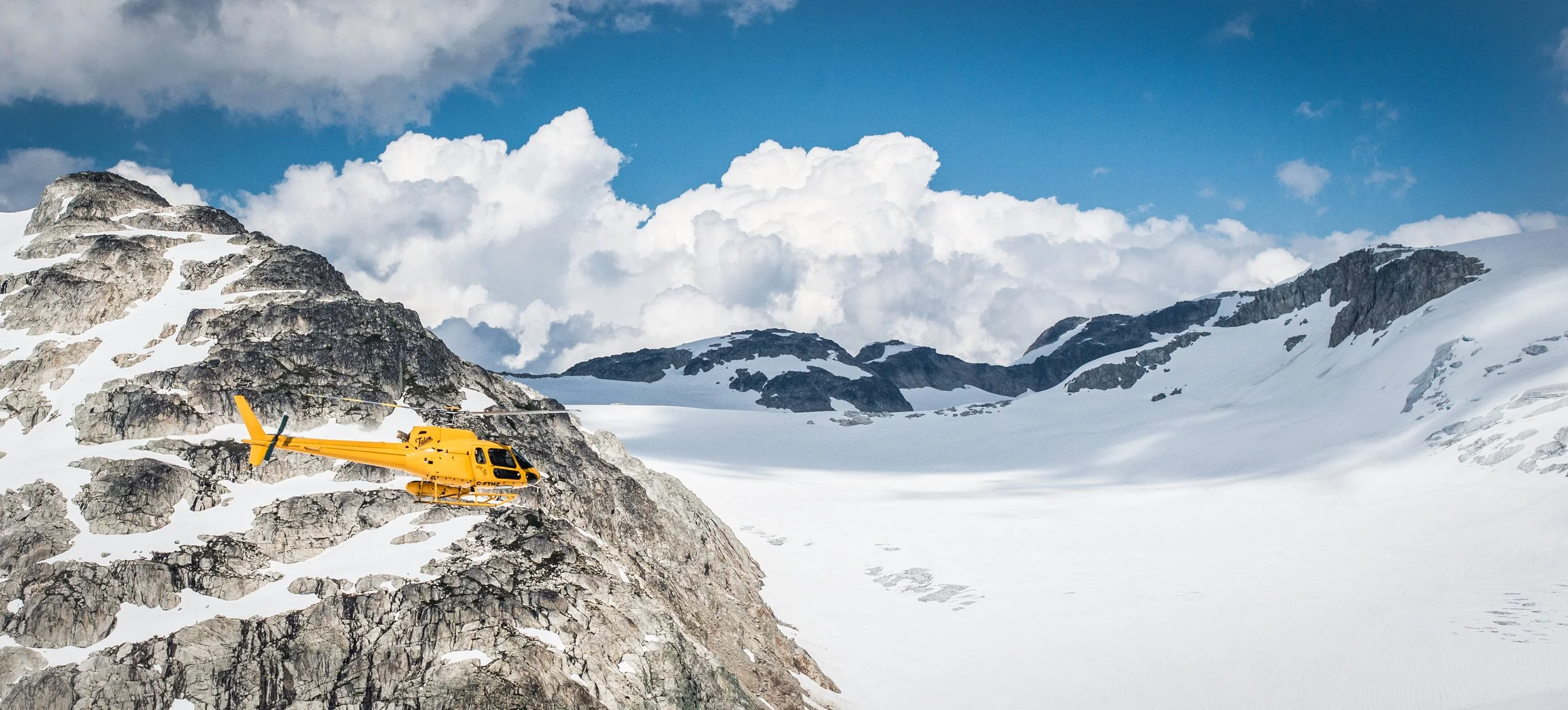 Yellow helicopter flying over snowy mountain landscape with rocky peaks and blue sky with clouds.