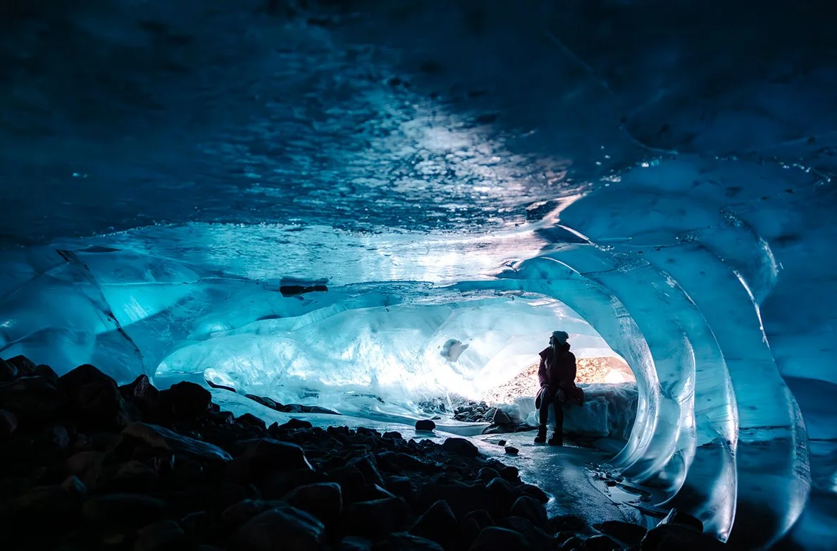 A person standing inside a blue, icy ice cave with smooth, curved walls and rocky floor, with sunlight coming through near the entrance.