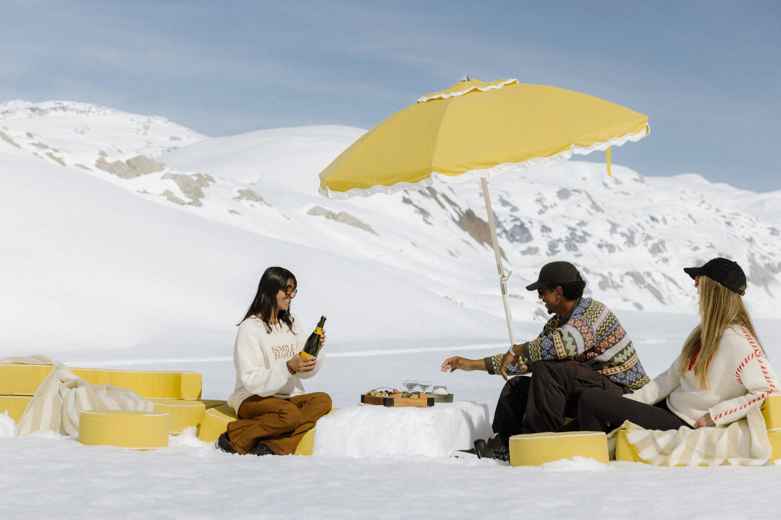 Group of people sitting on yellow benches outdoors in snowy landscape, with a yellow umbrella overhead. One person is holding a bottle, others are talking and enjoying themselves.
