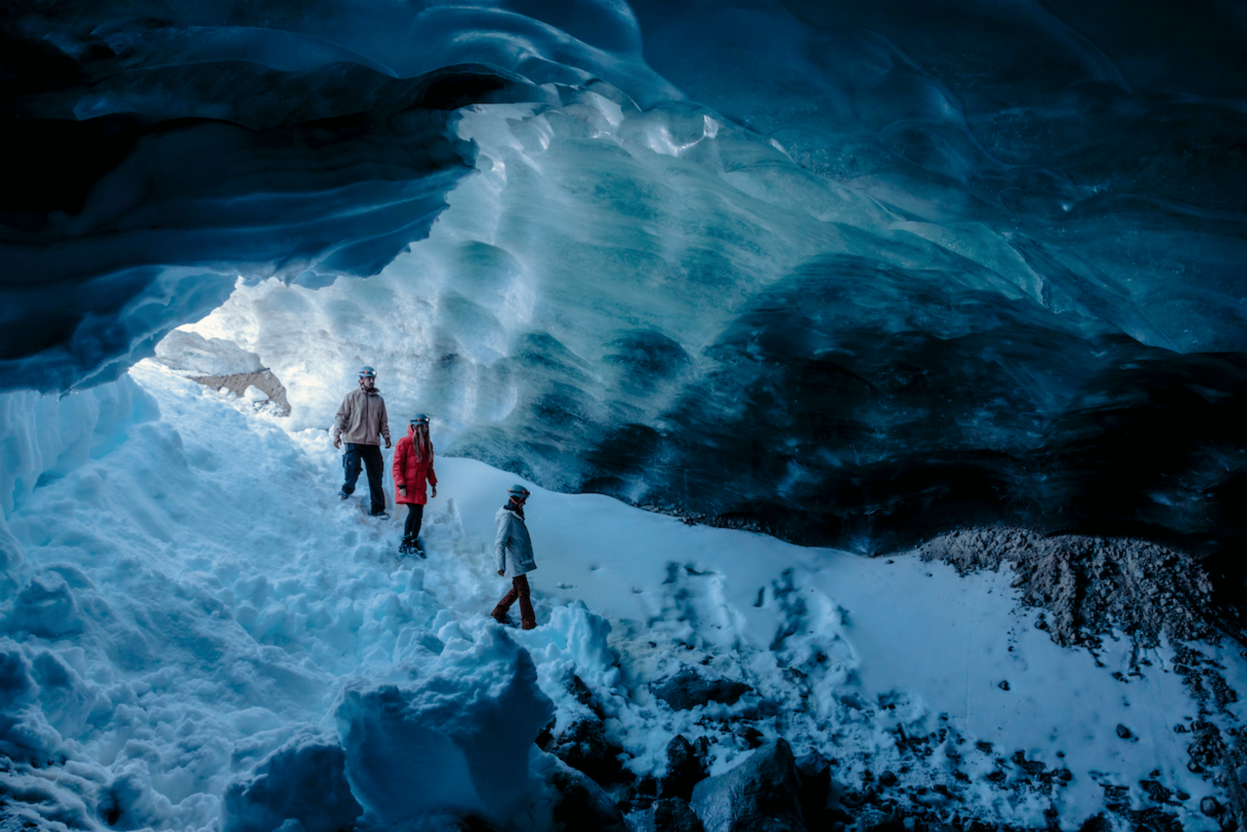 Group of four people in winter clothing and helmets exploring inside an ice cave with blue ice formations.