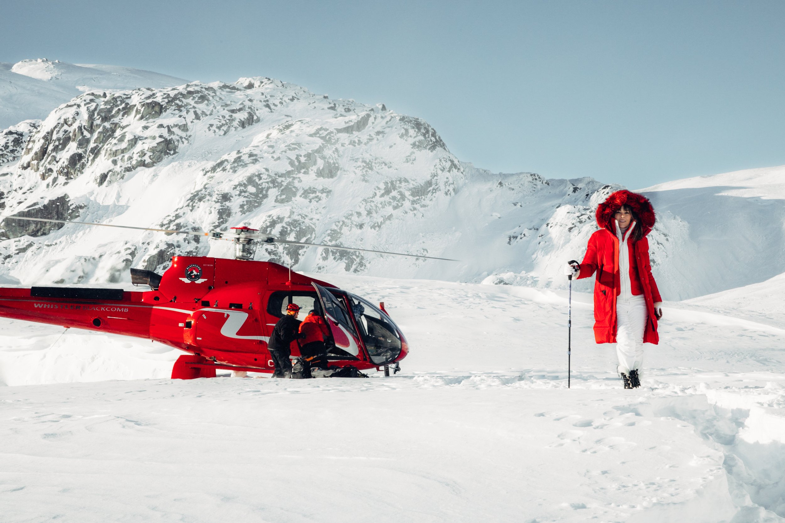 A woman in a red coat and white pants walks in the snow, holding a walking stick, with a helicopter and snowy mountains in the background.