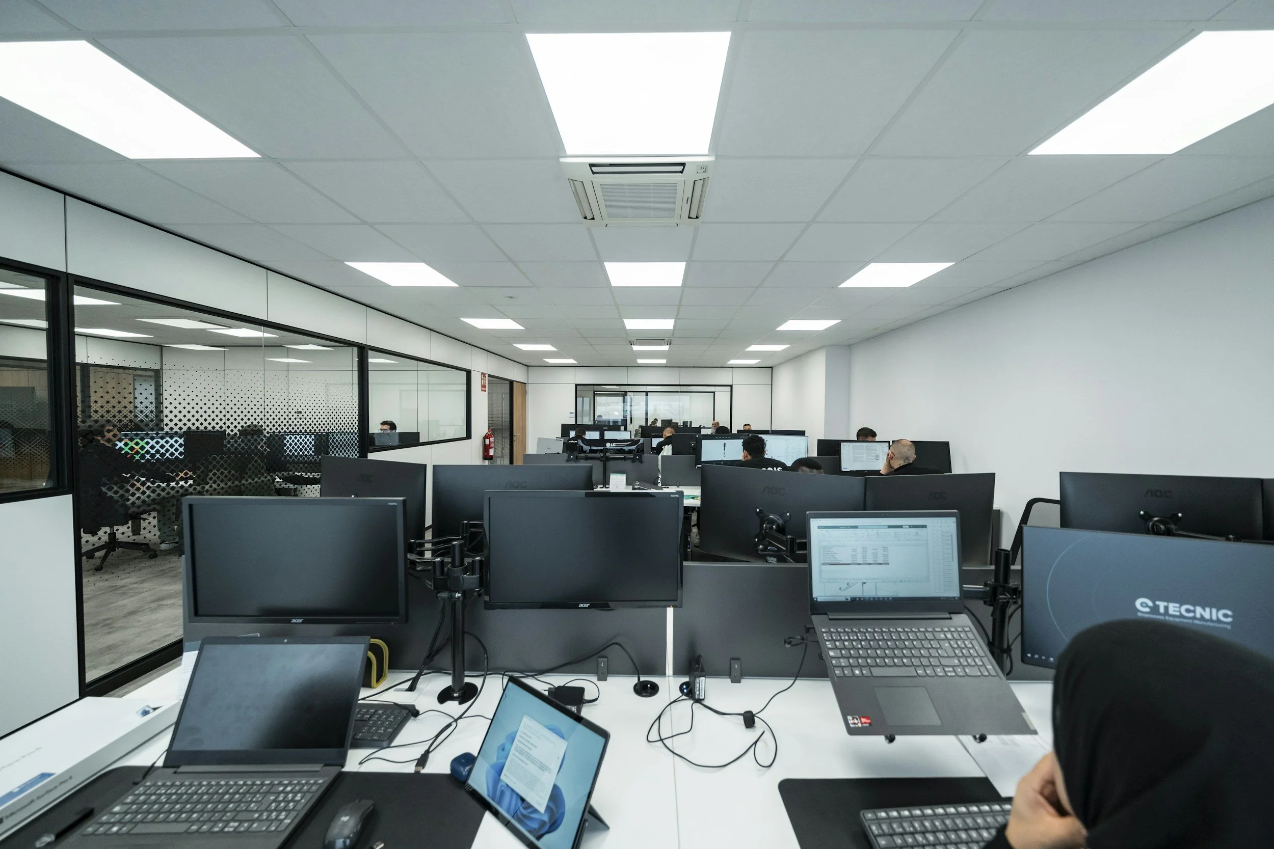 Office workspace with multiple desks, monitors, and people working at computers in a modern, well-lit office.