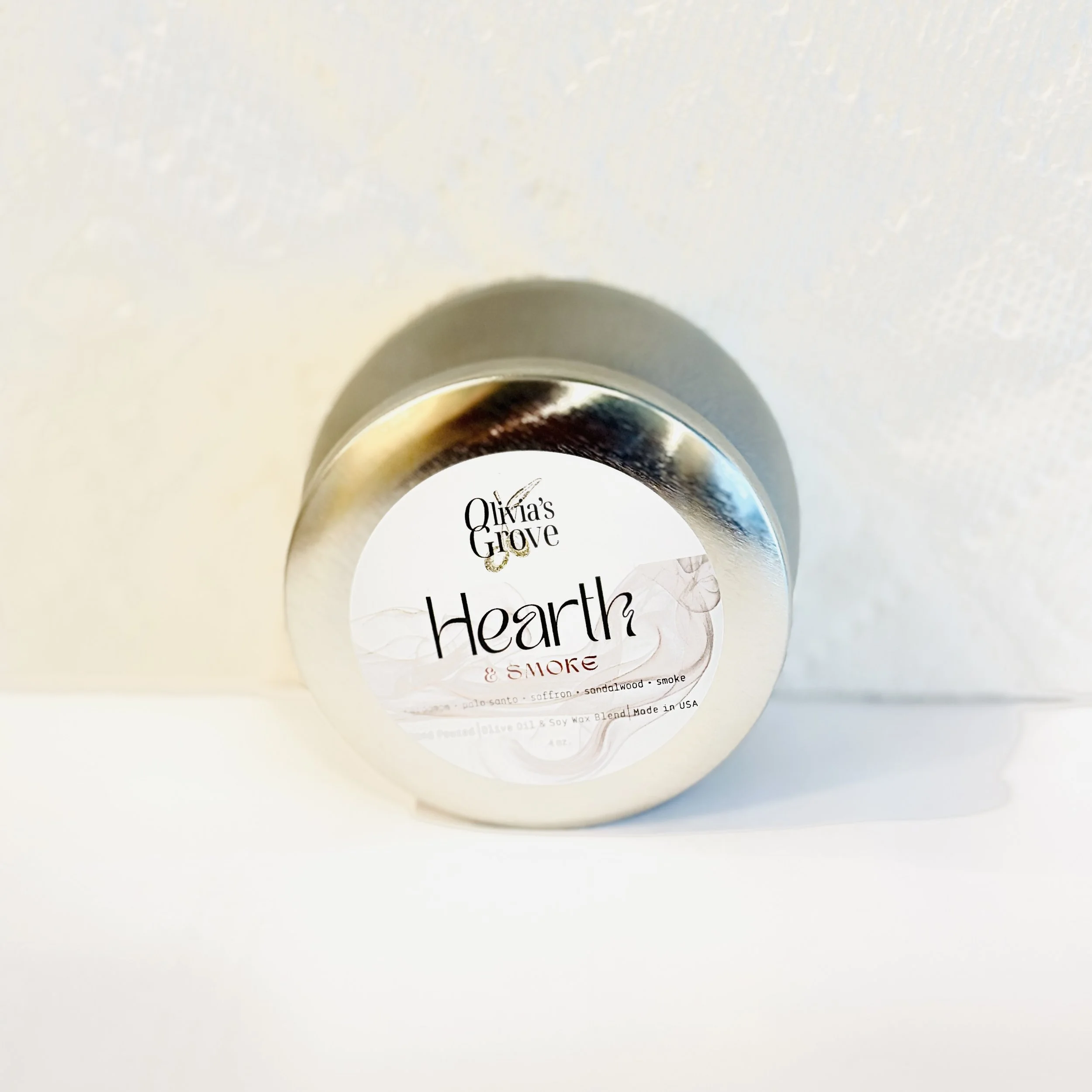 A tin jar labeled Olivia's Grove Hearth & Smoke, placed on a white surface against a textured white wall.