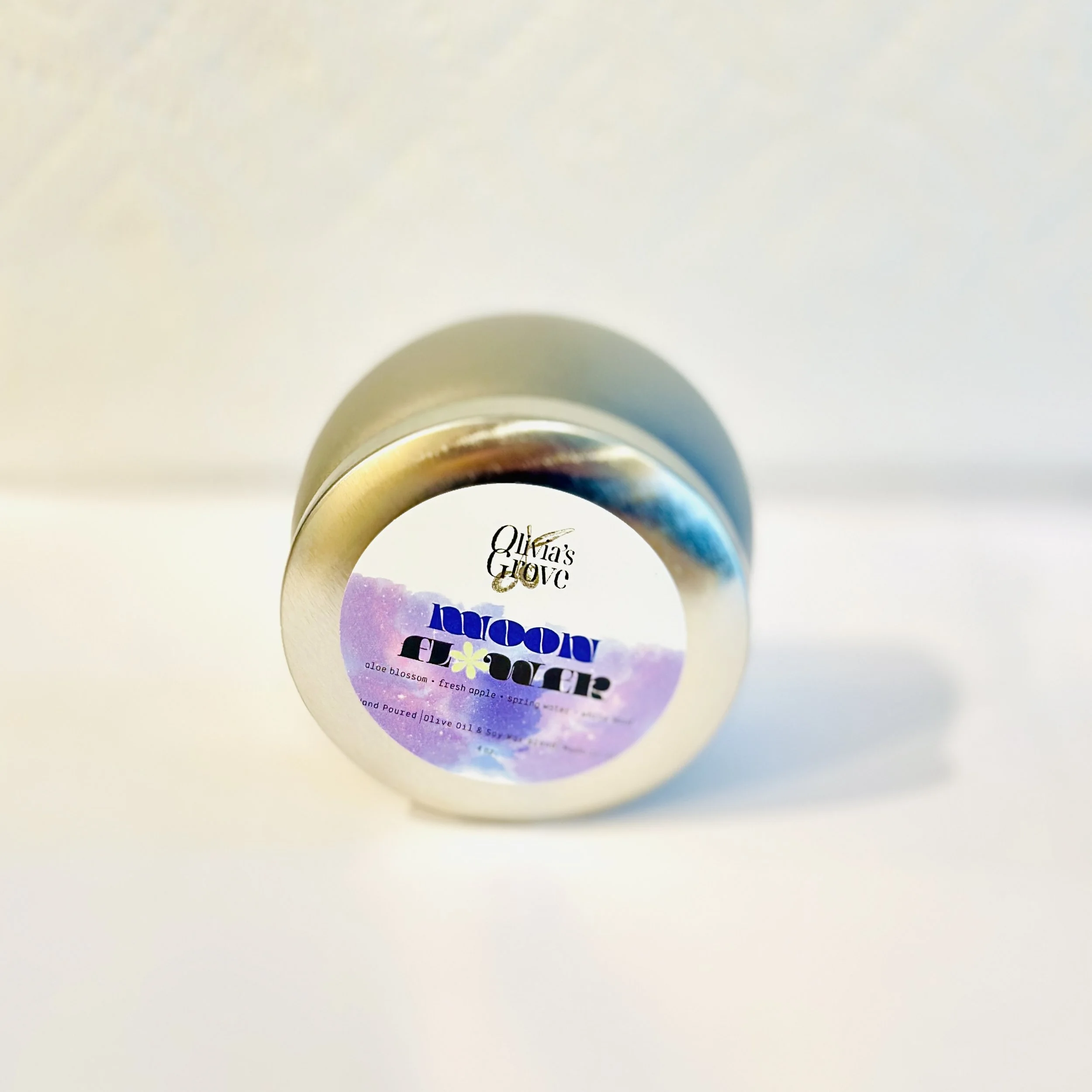 A small, metallic container with a label that reads "Olivia's Grove Moonflower," featuring a purple and blue watercolor design and text describing it as an aloe blossom, fresh apple, and spring.