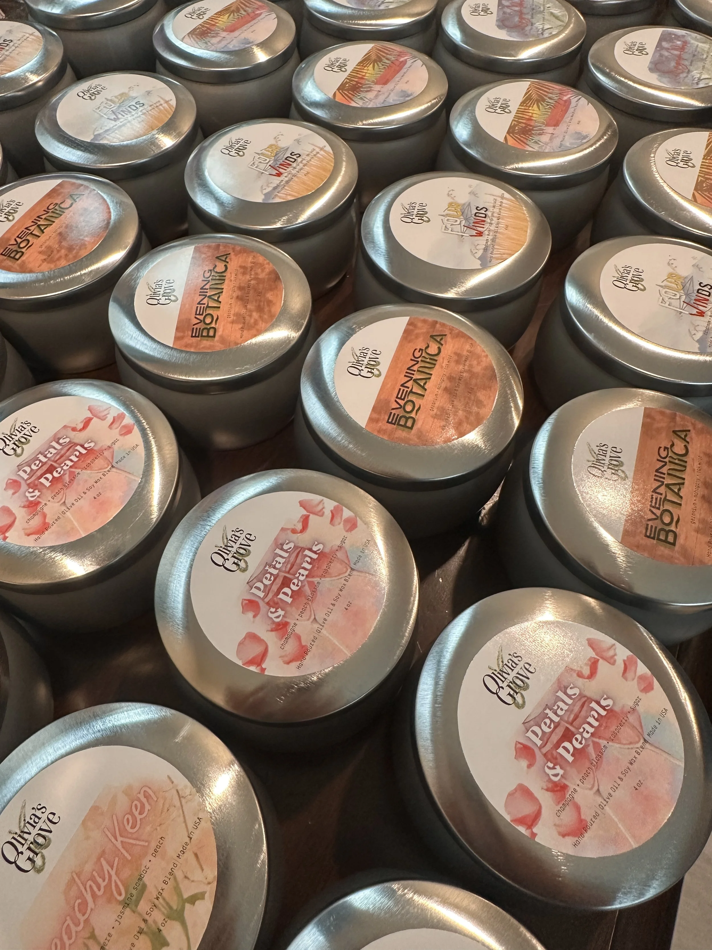 Multiple tins of homemade olive oil with labels featuring different botanical and floral designs, some with titles like 'Dear Peals & Pearls' and others with 'Evening Botanic'.