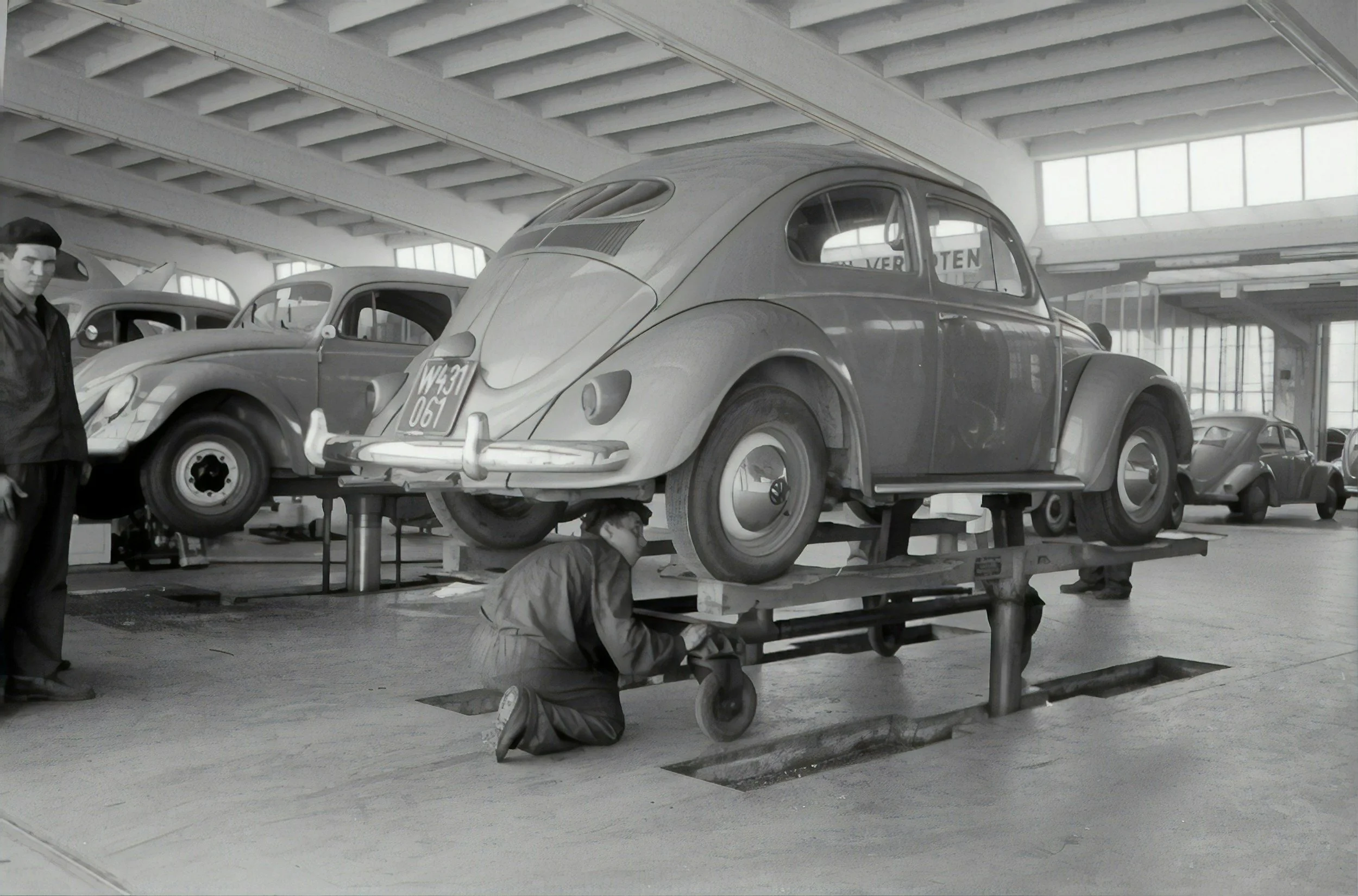 Black and white photo of a vintage car factory, with a worker inspecting a classic car elevated on a platform, surrounded by other vintage cars.