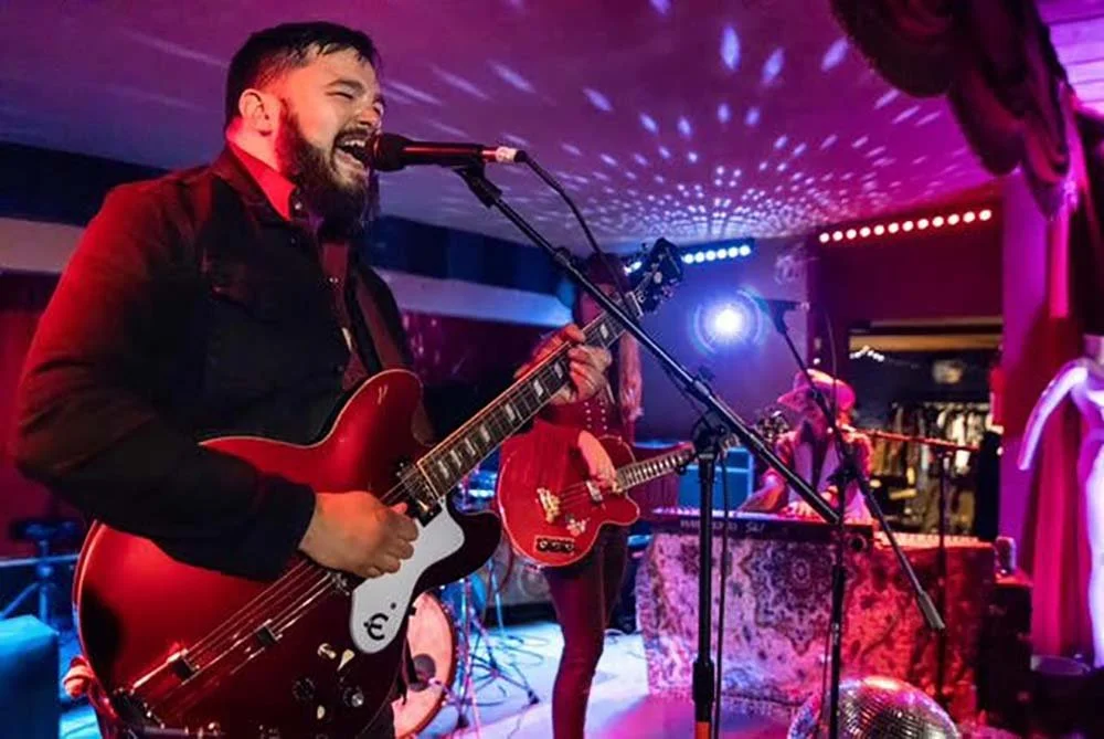 Musicians performing live on stage with guitars and microphone, colorful stage lighting, and a patterned tablecloth in the background.
