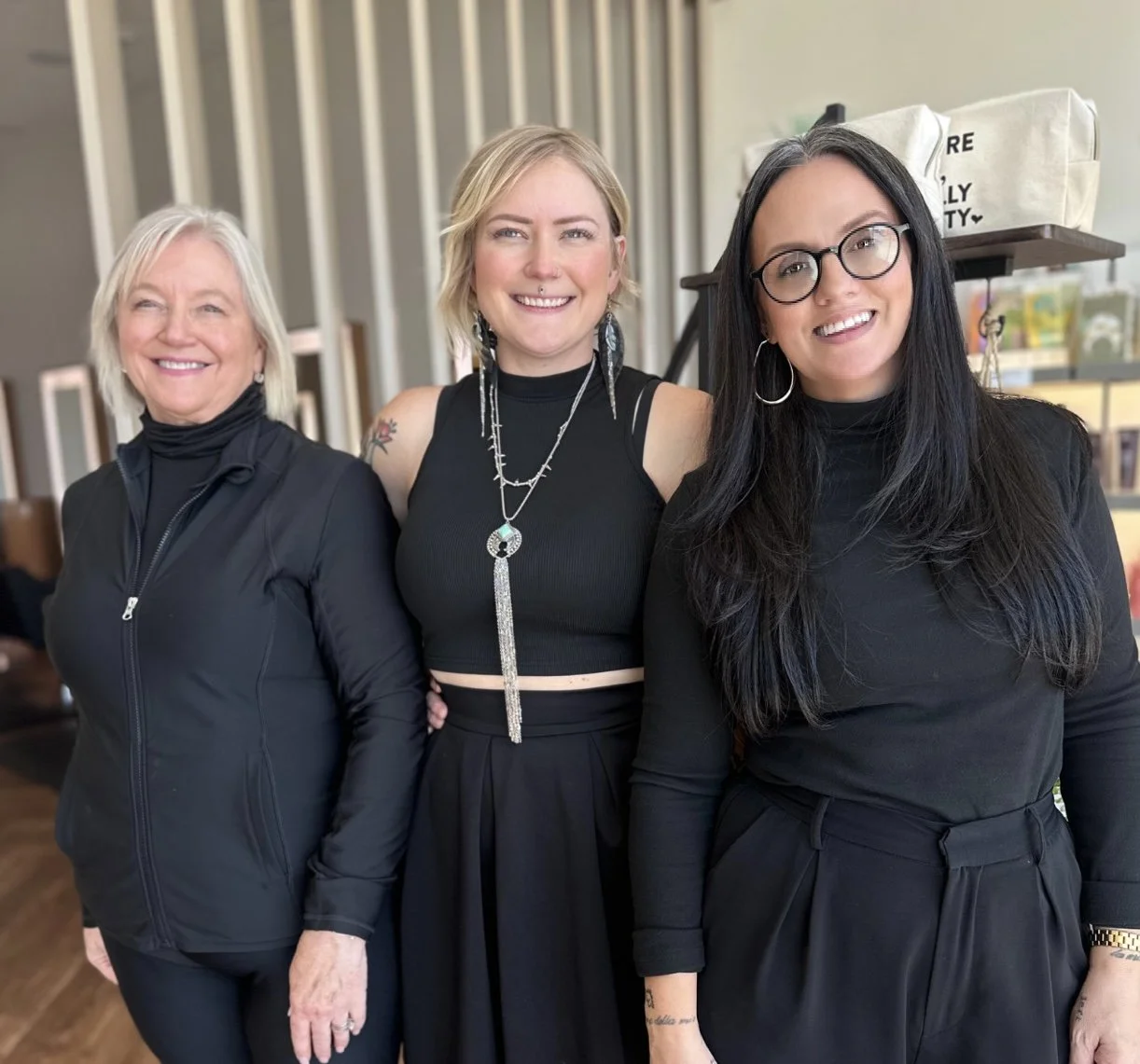 Three women standing together indoors, smiling, dressed in black clothing.