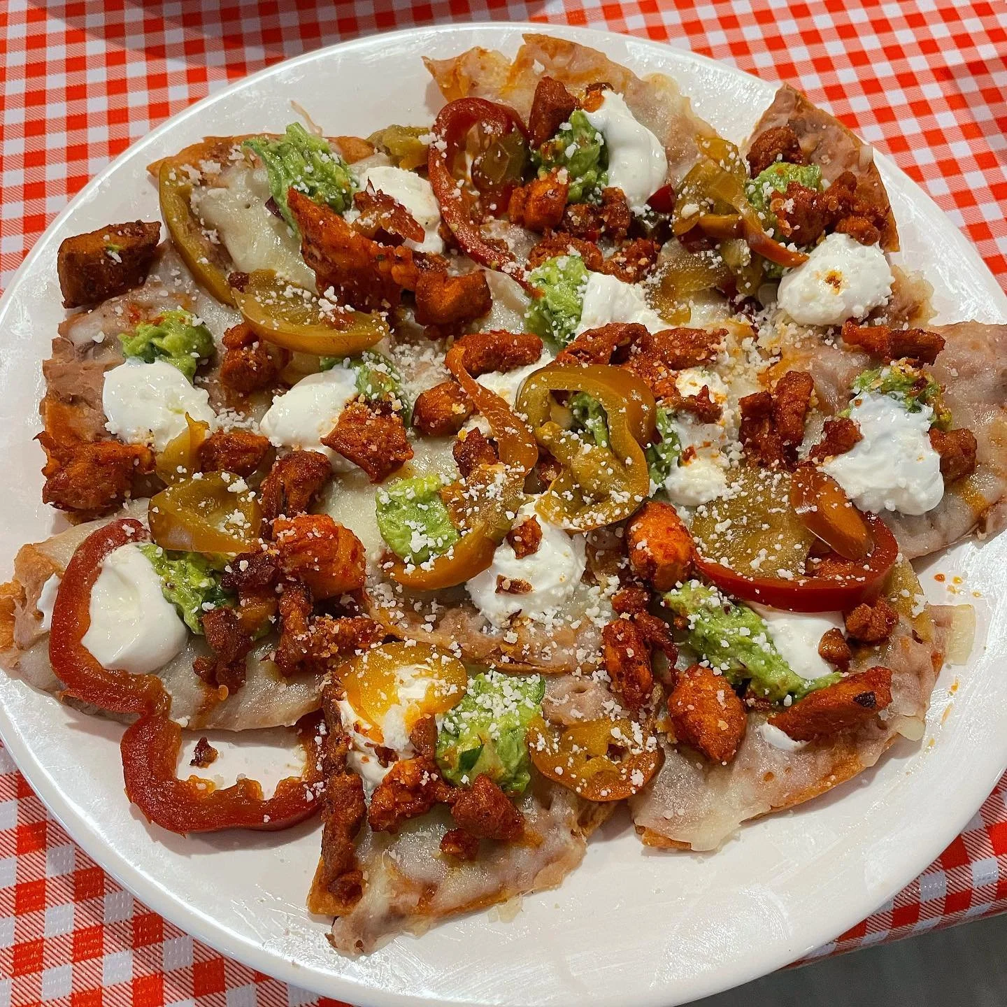 Pizza with bell peppers, diced chicken, dollops of cream, cheese, and lettuce on top, served on a white plate with a red and white checkered tablecloth.