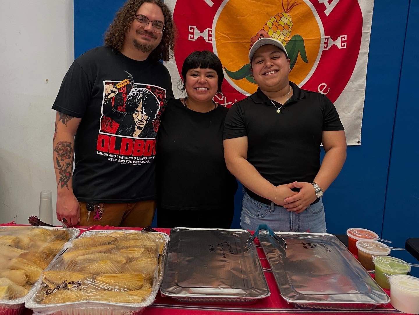 Three people standing behind a table with food and containers. They are smiling, with a man on the left with curly hair, glasses, tattoos, and a black "Oldboy" graphic T-shirt, a woman in the middle with short black hair, and a man on the right with a cap, black shirt, and light jeans. They are in front of a red and yellow banner with a corn graphic.