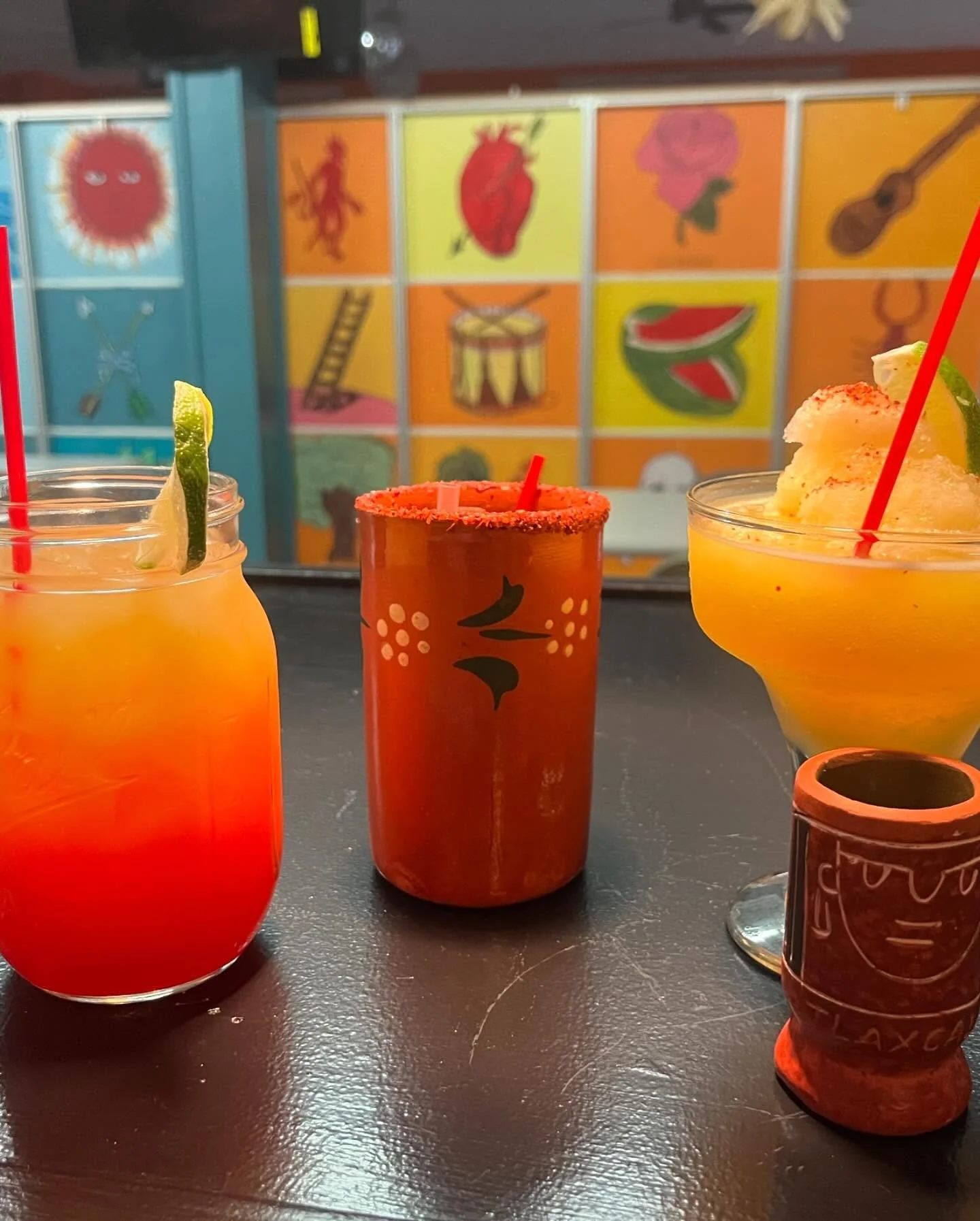 Three colorful cocktails with garnishes on a dark surface in front of a bright mural with colorful images of traditional Mexican symbols.