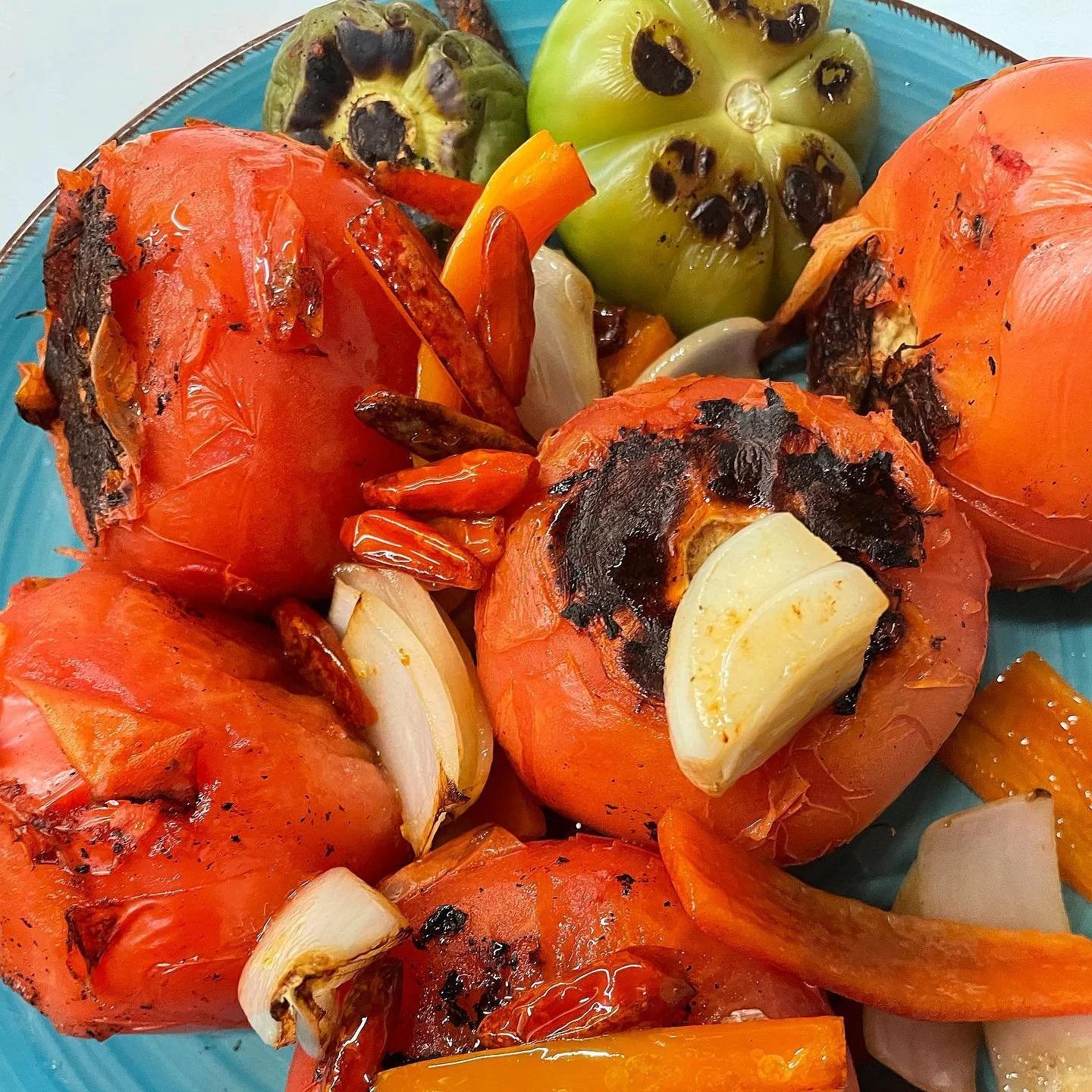 Charred tomatoes with garlic cloves and sliced onions, served with roasted peppers and a few cherry tomatoes with black spots.